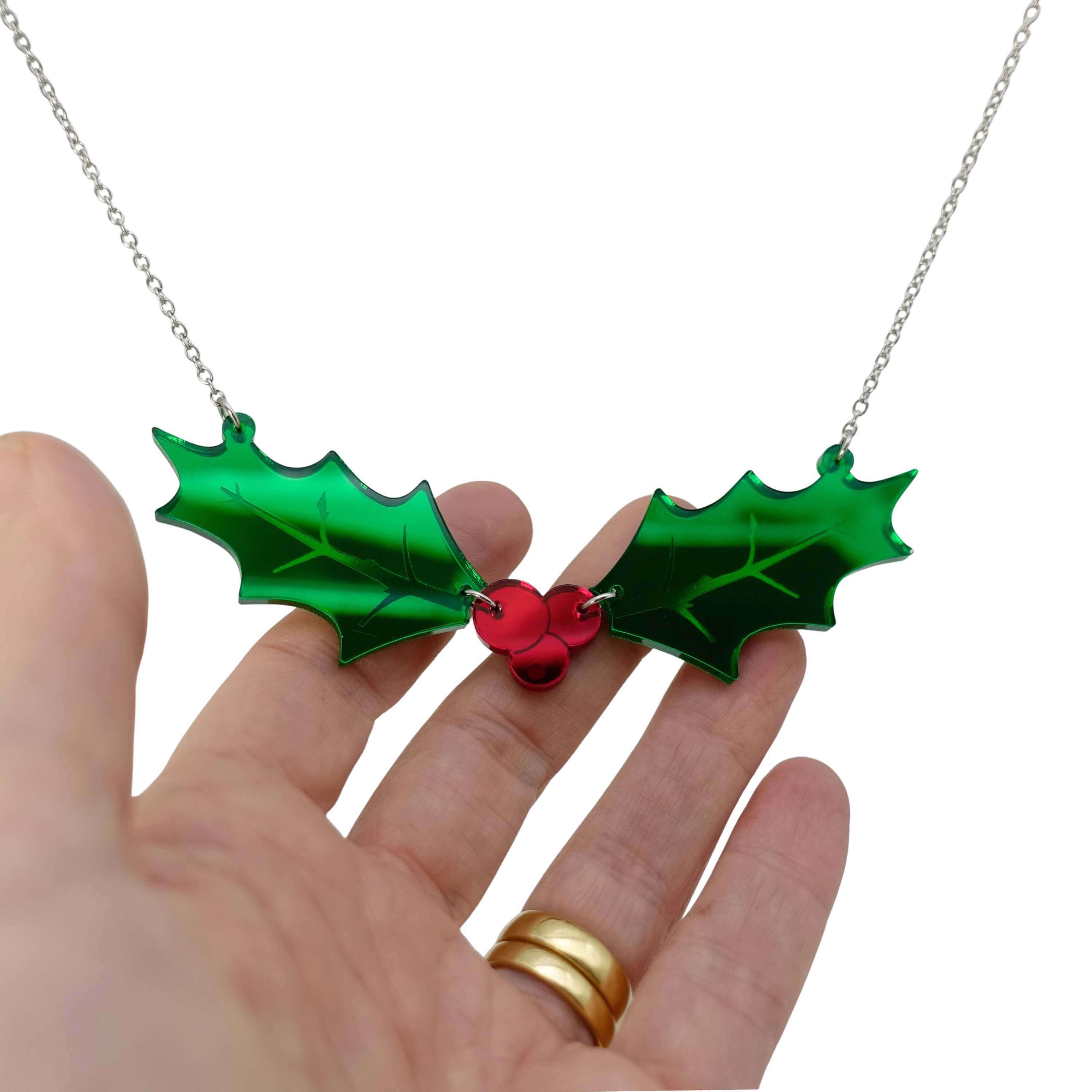 Naturalist Holly necklace with two holly leavs joined by a trio of red berries in mirror acrylic, shown hanging on a stainless steel chain with my hand behind for scale and to show the reflectiveness of the acrylic. 