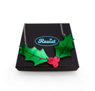 A naturalist Holly necklace with two holly leavs joined by a trio of red berries in mirror acrylic, shown on a Wear and Resist gift box. 