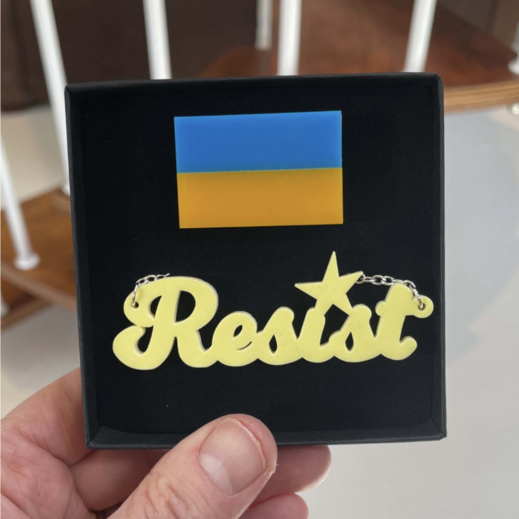 An I stand with Ukraine pin shown with a Resist necklace. Designed by Sarah Day for Wear and Resist. 