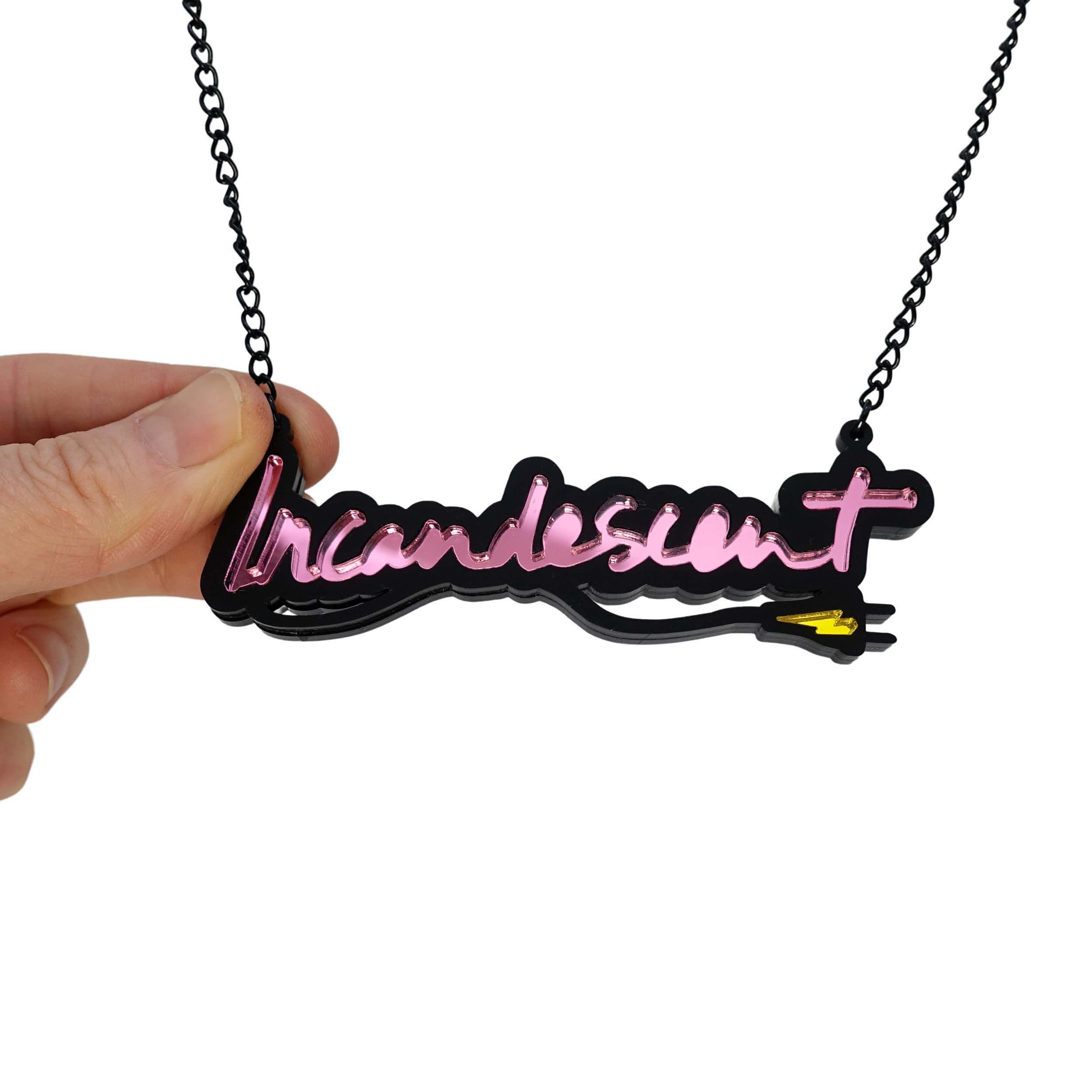 Incandescent neon necklace in pink with a yellow lightning bolt plug, on a black chain adjustable to three lengths. Shown hanging against a white backround with my hand holding the chain for scale. 