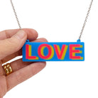 LOVE necklace in colourful blue, hot pink and yellow, shown hanging on a stainless steel chain with my hand holding it for scale. 