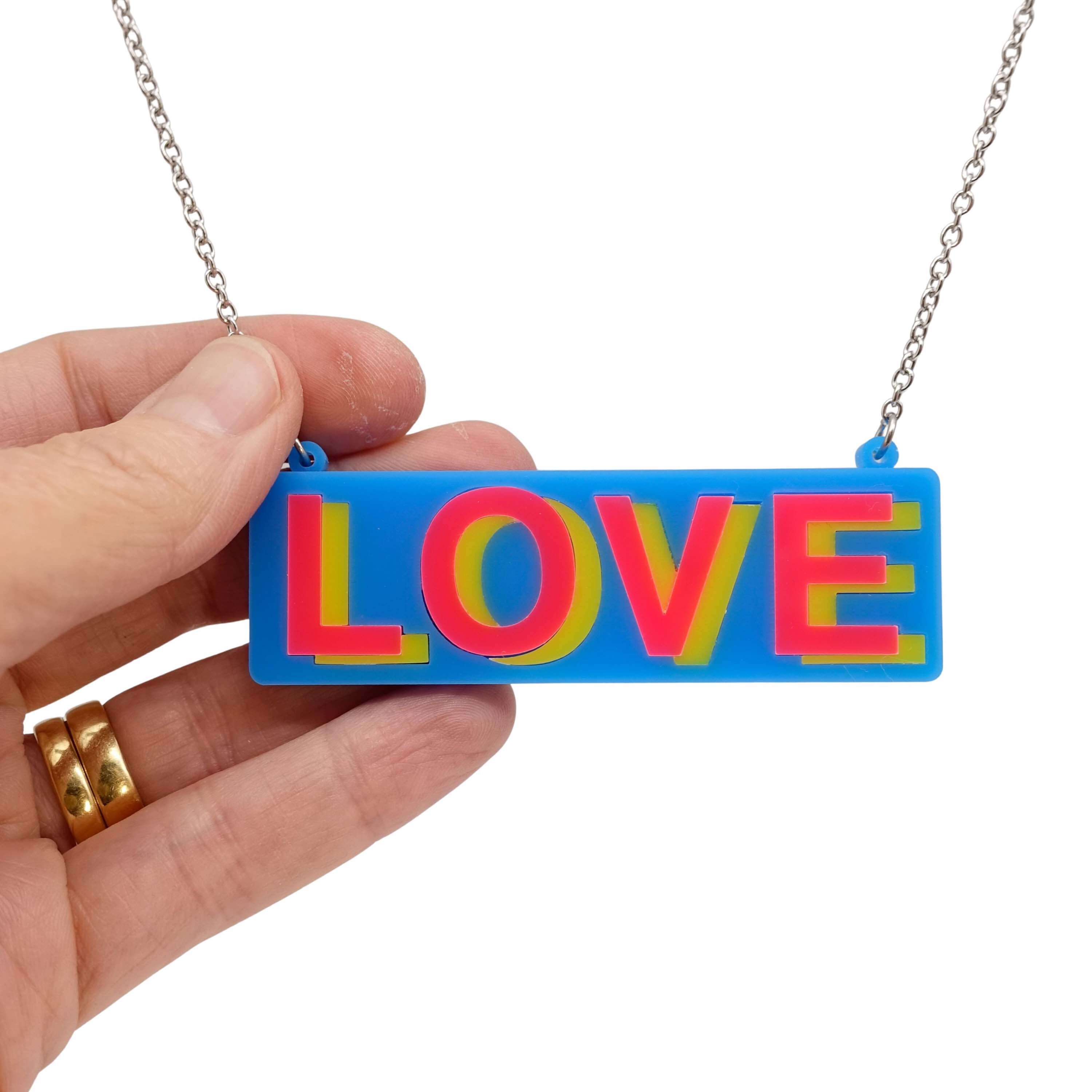 LOVE necklace in colourful blue, hot pink and yellow, shown hanging on a stainless steel chain with my hand holding it for scale. 