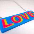 A close up of the LOVE necklace in colourful blue, hot pink and yellow, shown laying on a white surface.