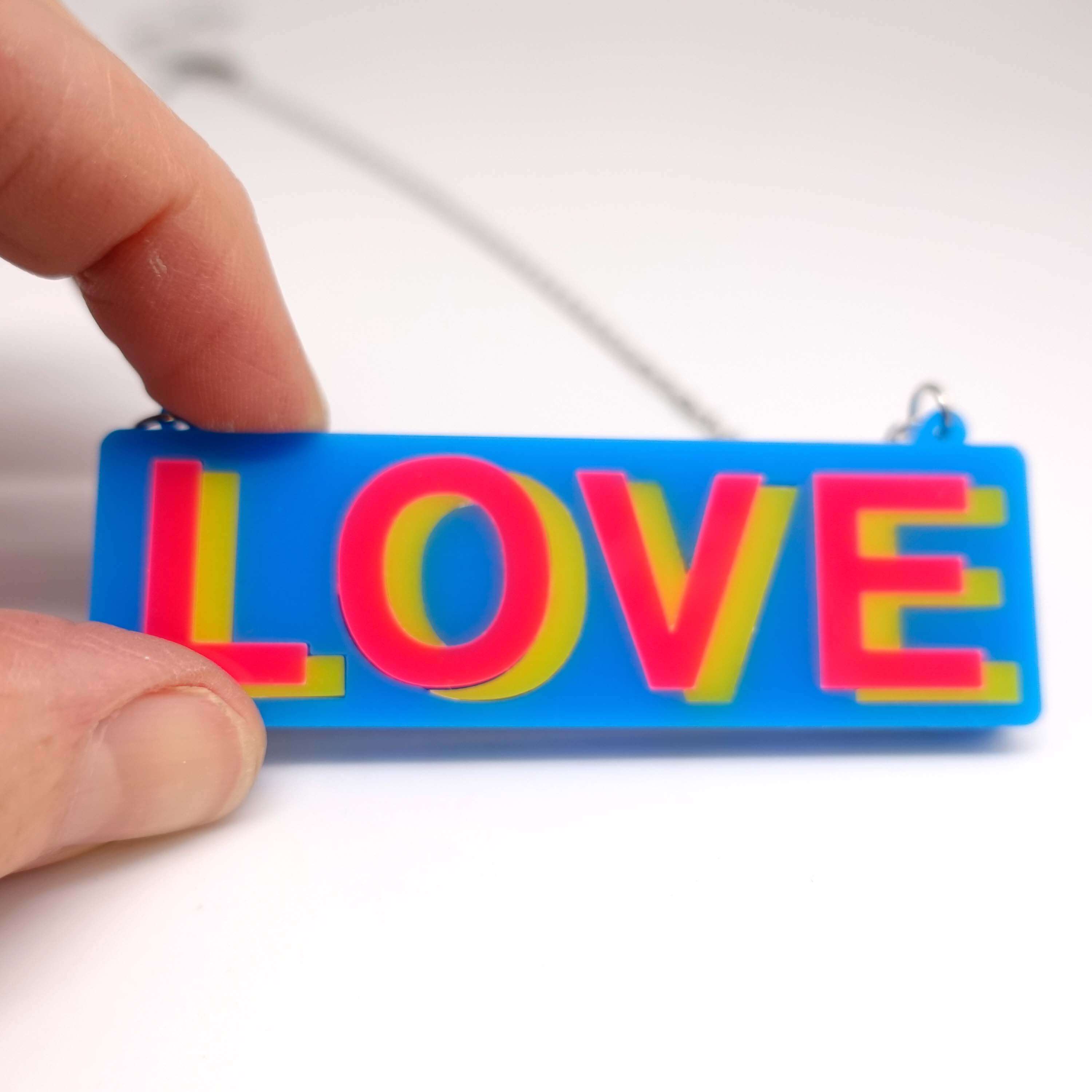 A close up of the LOVE necklace in colourful blue, hot pink and yellow, shown held against a white surface.