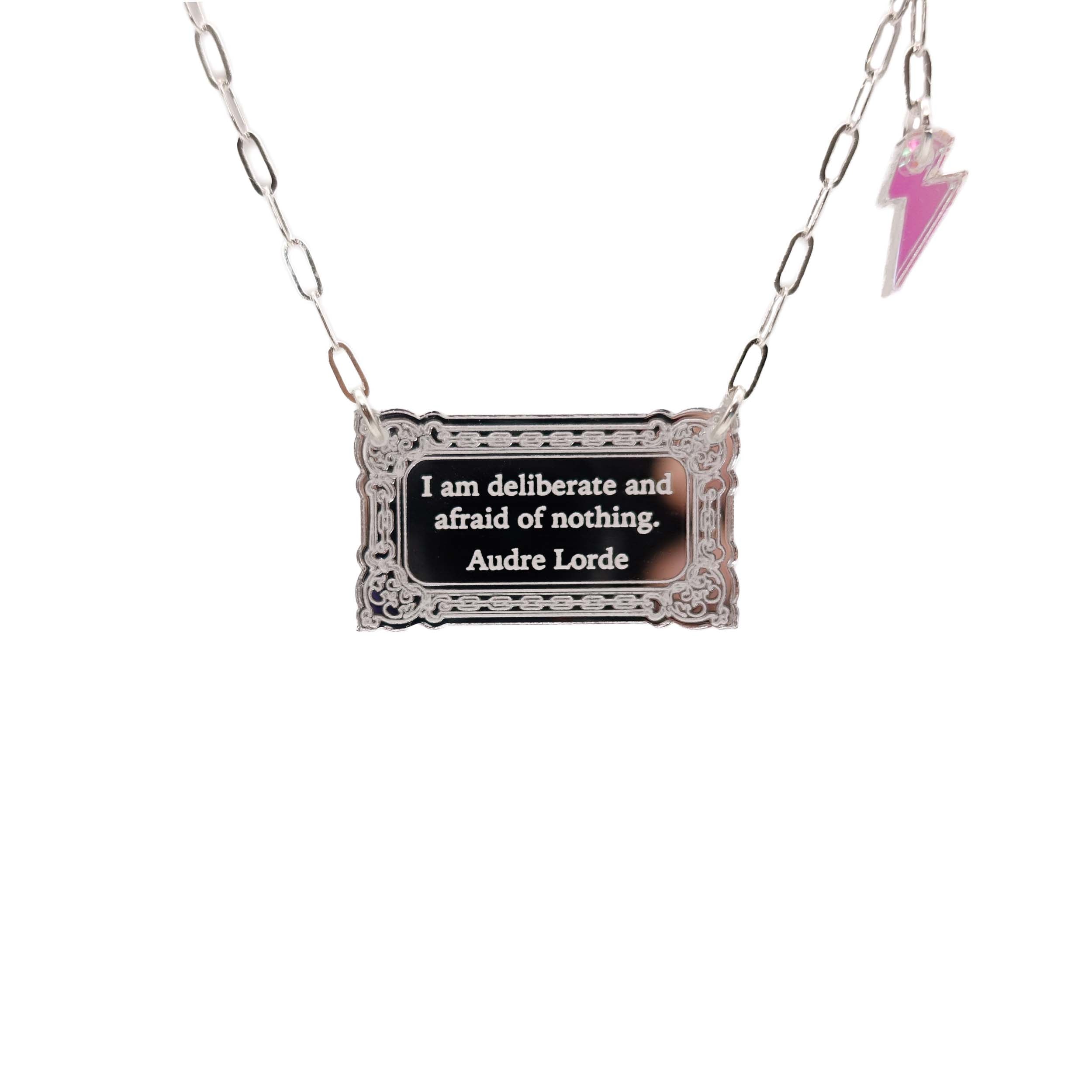 Literary Quote necklace etched with a quote by Audre Lorde, 'I am deliberate and afraid of nothing.' Shown hanging in front of a white background with a small iridescent lightning bolt at the end of the silver paperclip chain. 