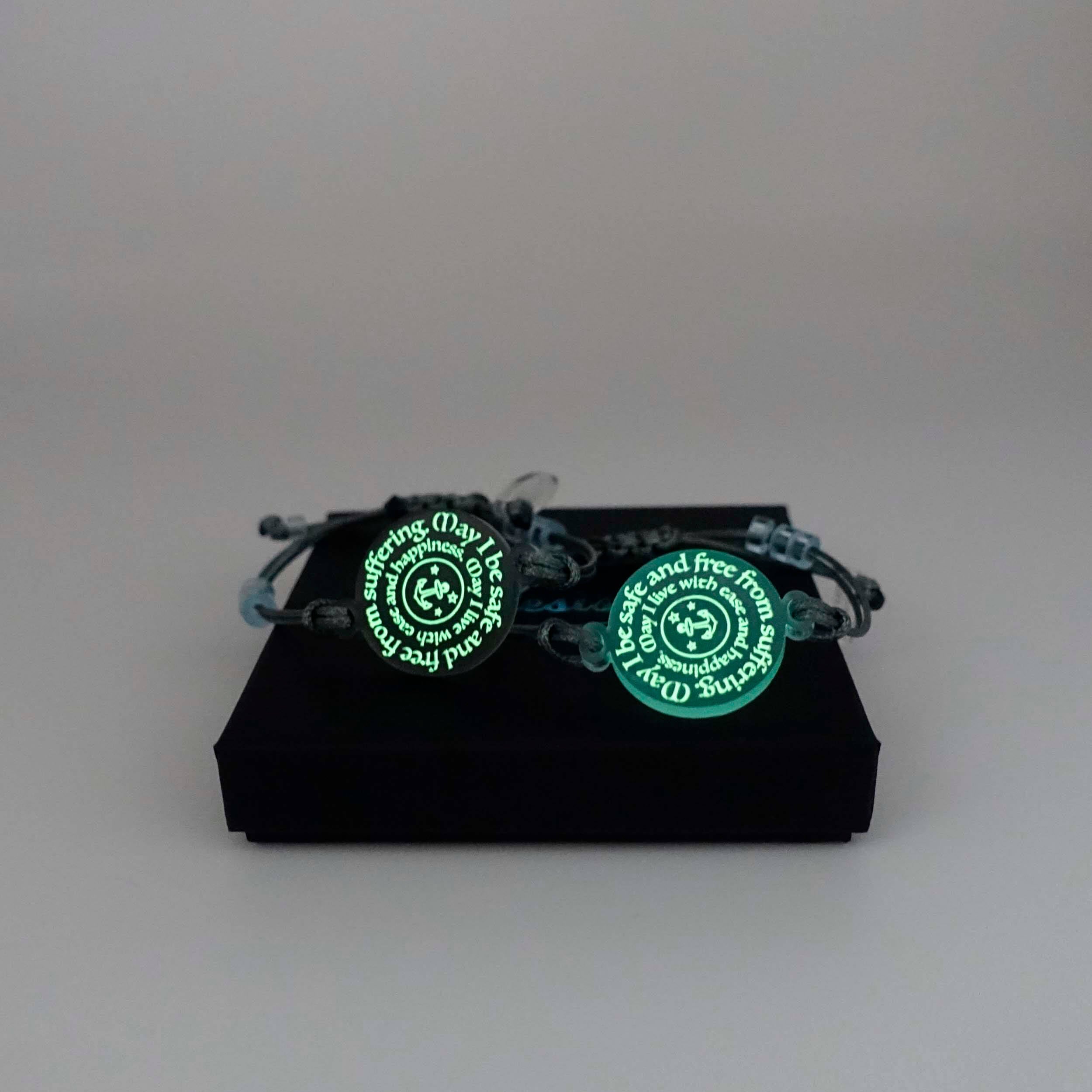 May I Be Safe bracelets in sea foam and shale colours shown glowing in the dark on a Wear and Resist gift box. 