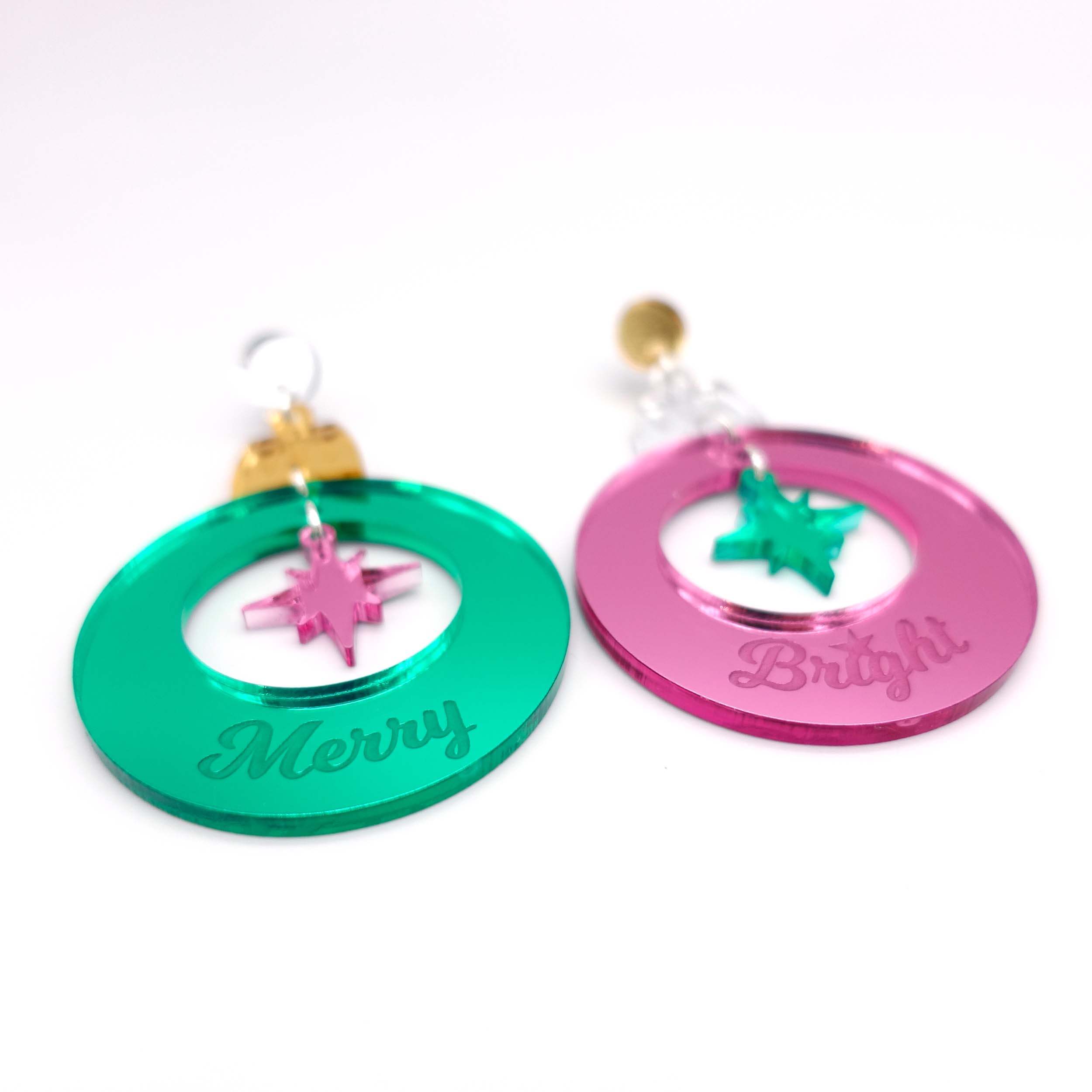 Pink and green Merry & Bright Christmas bauble earrings shown laying on white surface. 