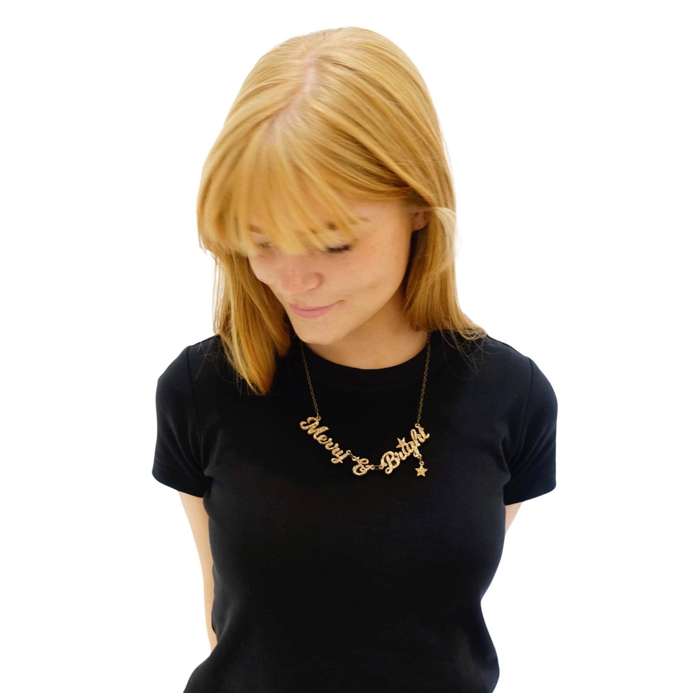 Eliza wears a gold glitter Merry & Bright necklace against a black t-shirt. 