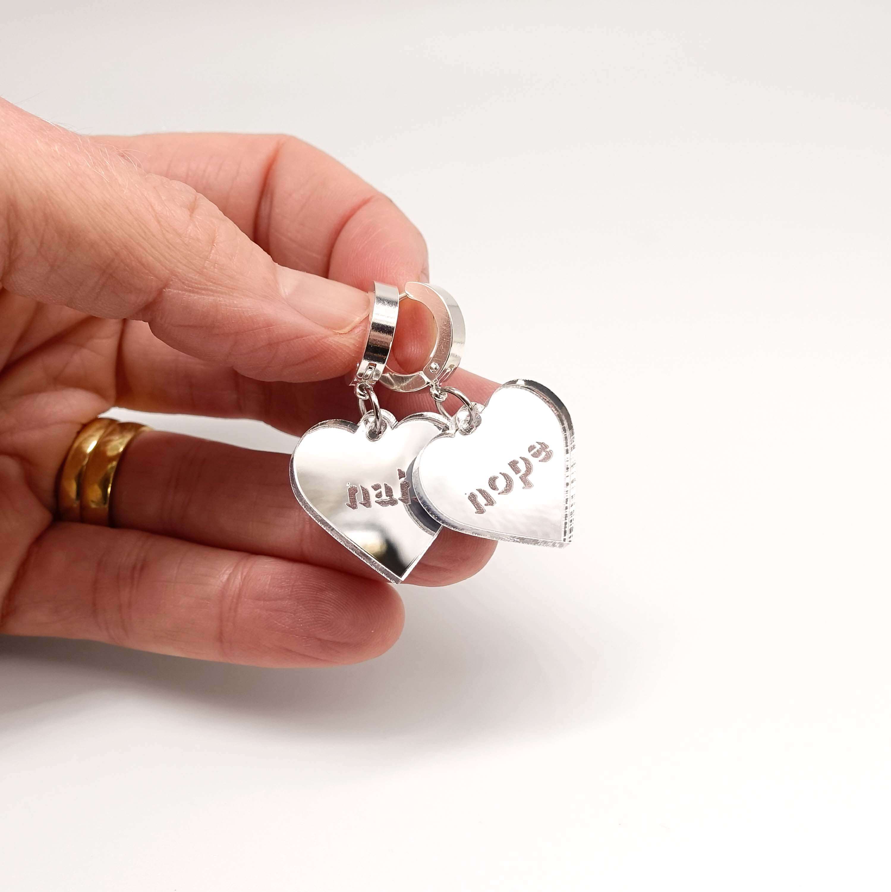 Nah Nope heart shaped huggie hoop earrings in silver mirror shown held in my hand with a white surface behind. 