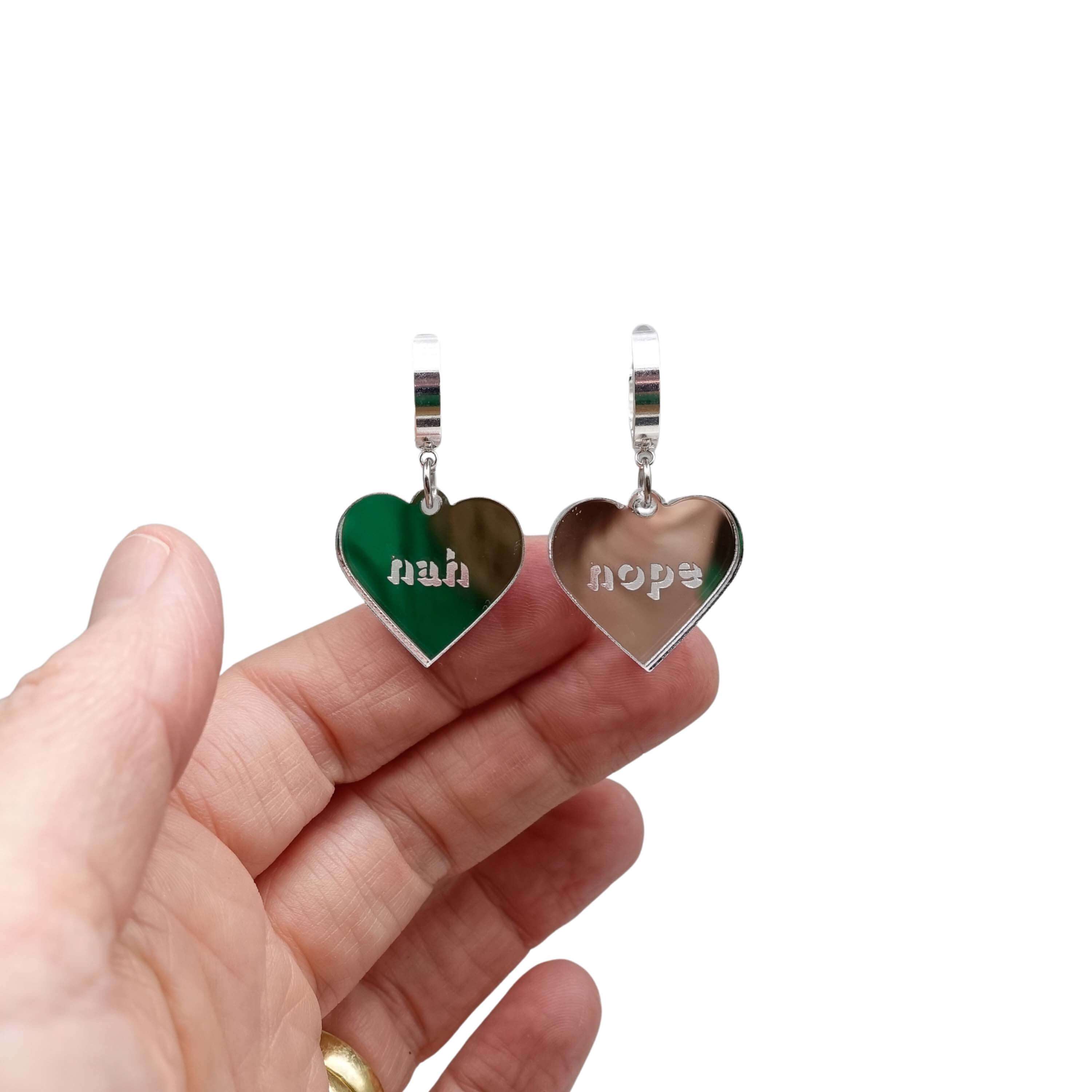 Nah Nope heart shaped huggie hoop earrings in silver mirror shown hanging against a white background with my hand behind for scale. 
