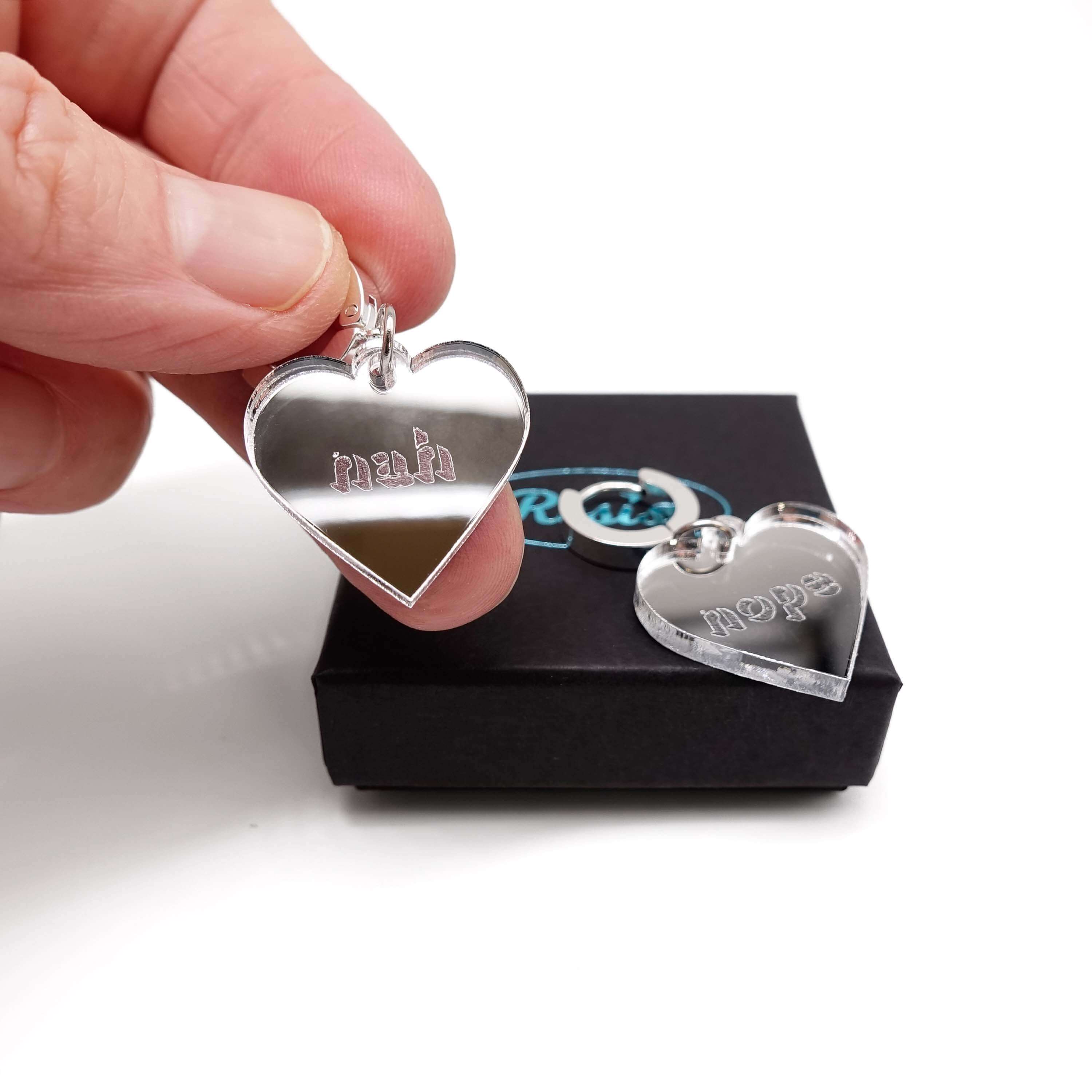 Nah Nope heart shaped huggie hoop earrings in silver mirror, the Nope is laying on the Wear and Resist gift box and I am holding the Nah up. 