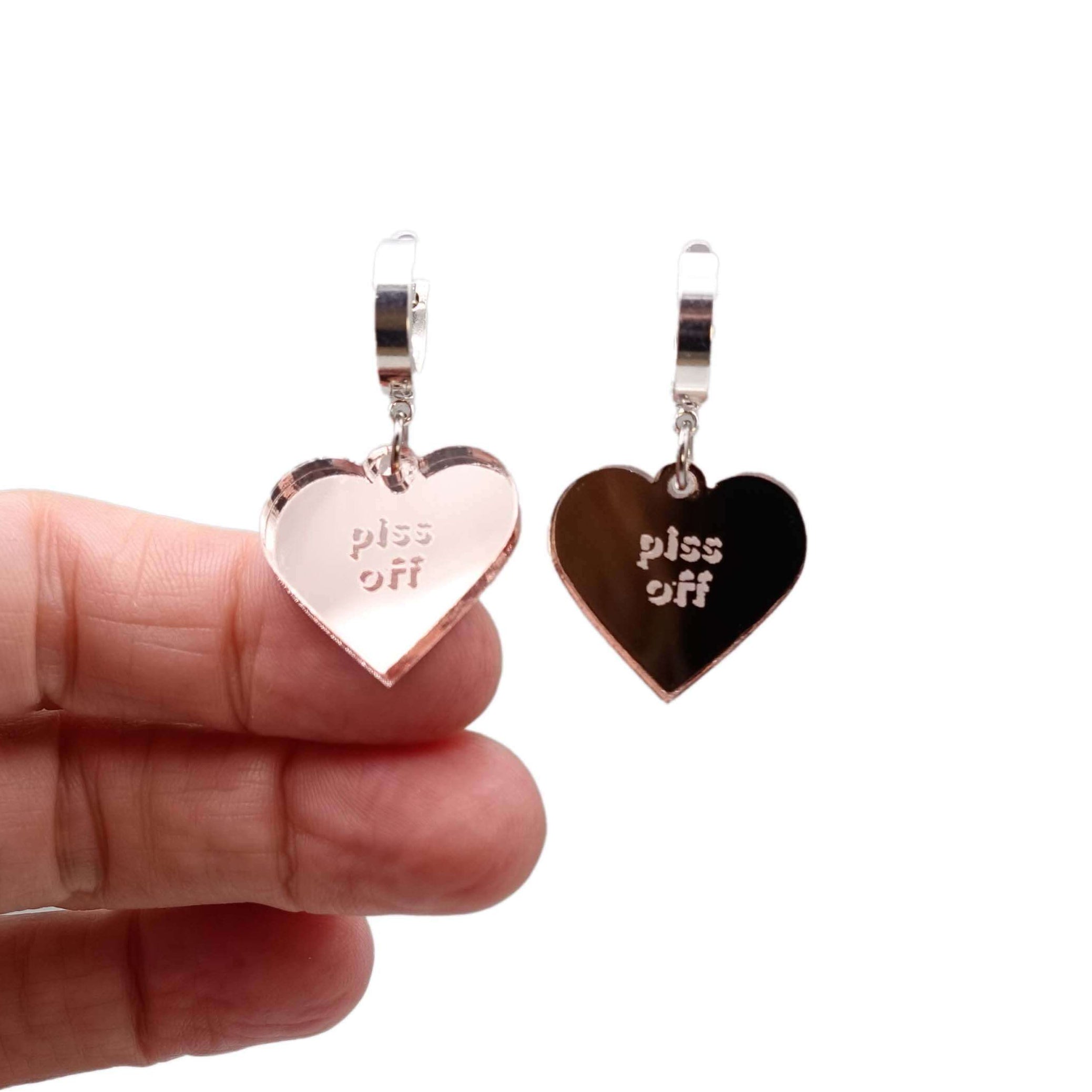 Heart-shaped earrings with 'piss off' engraved on them held by a hand on a white background