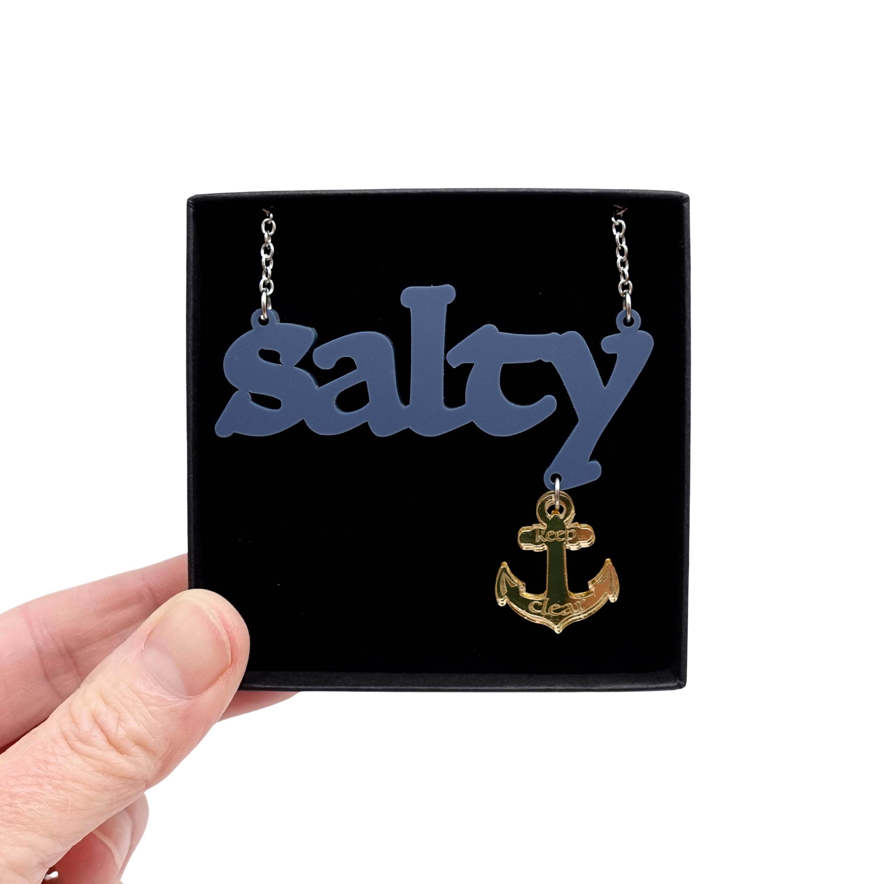 Salty necklace with an etched anchor that says 'keep clear' shown on a stainless steel chain held up in a Wear and Resist gift box. 