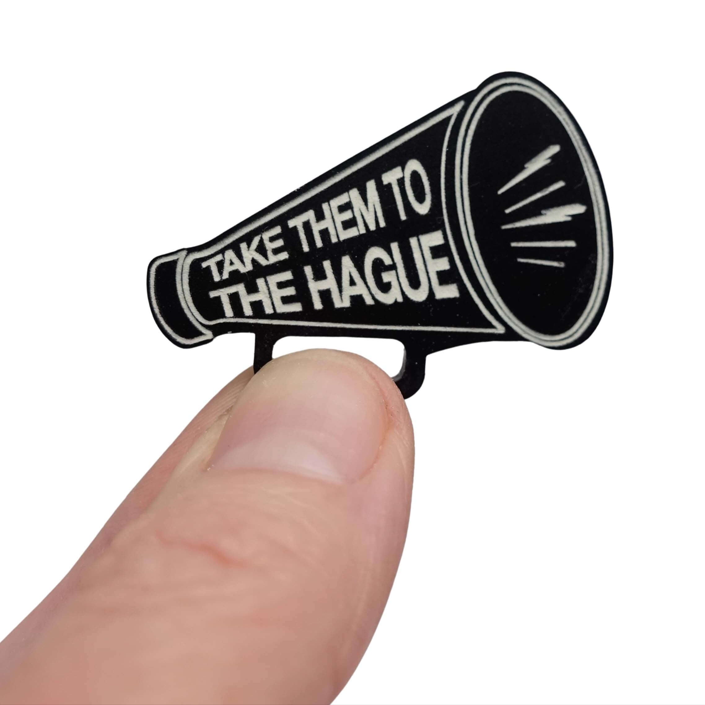 Small megaphone brooch etched with the words Take them to the Hague in glow-in-the-dark-pigment, shown held up in my fingers against a white background. 