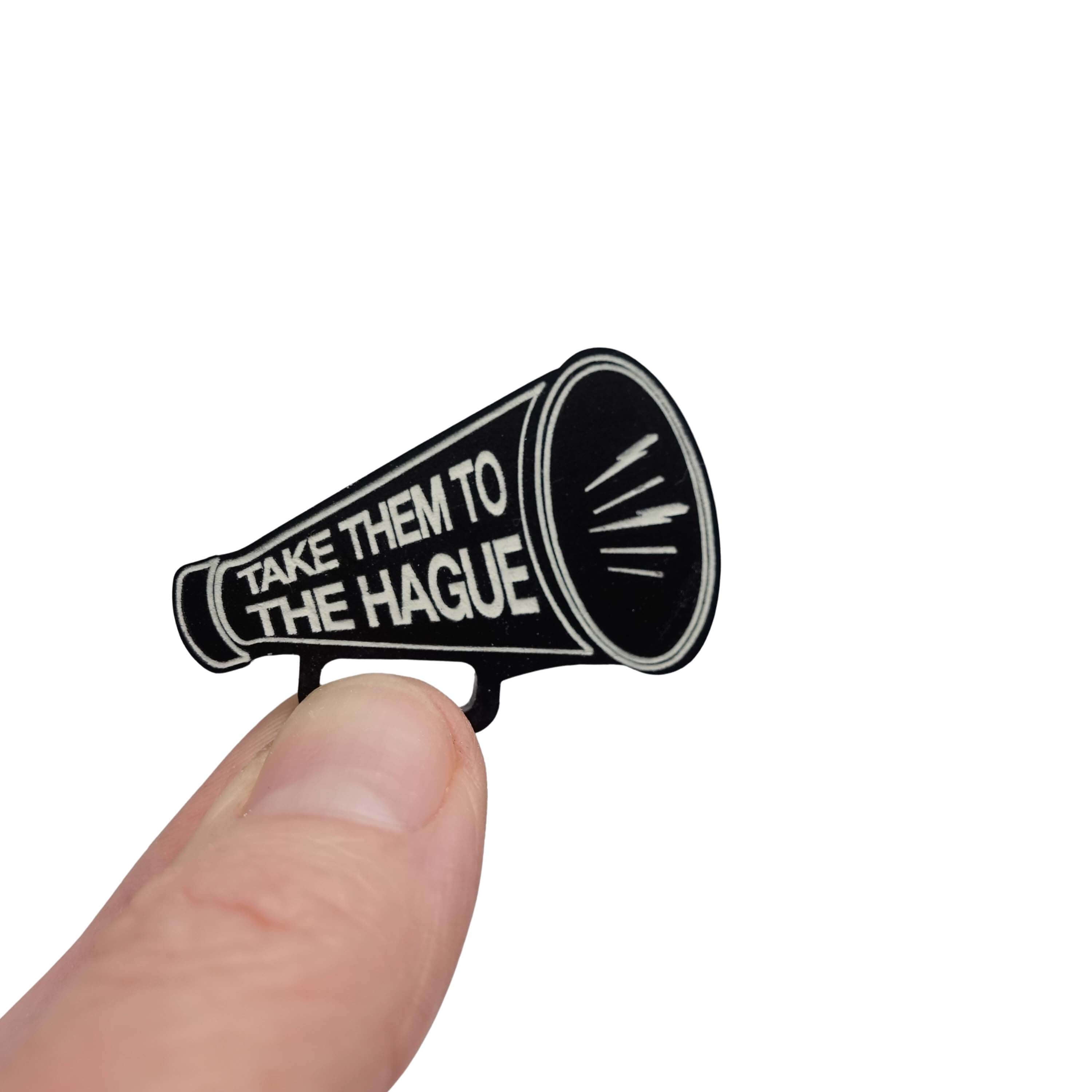 Small megaphone brooch etched with the words Take them to the Hague in glow-in-the-dark-pigment, shown held up in my fingers against a white background. 