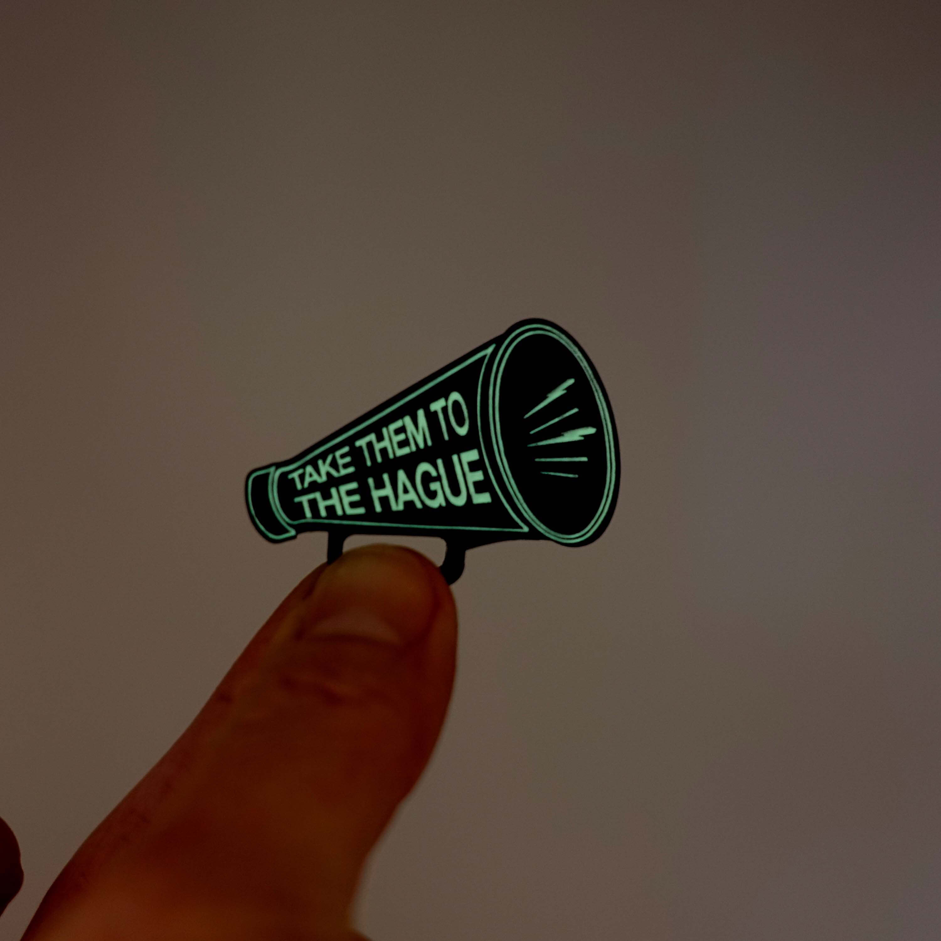 Small megaphone brooch etched with the words Take them to the Hague in glow-in-the-dark-pigment, shown held up in my fingers glowing in the dark. 