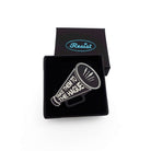 Small megaphone brooch etched with the words Take them to the Hague in glow-in-the-dark-pigment, shown in a small Wear and Resist gift box. 