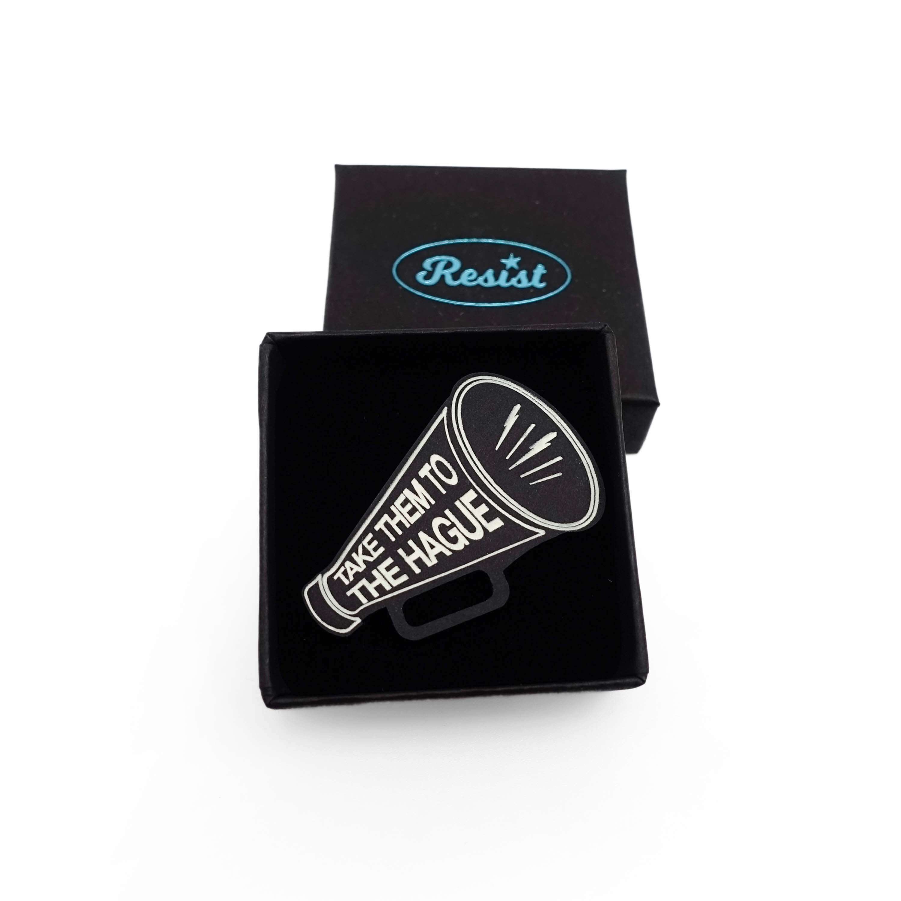 Small megaphone brooch etched with the words Take them to the Hague in glow-in-the-dark-pigment, shown in a small Wear and Resist gift box. 