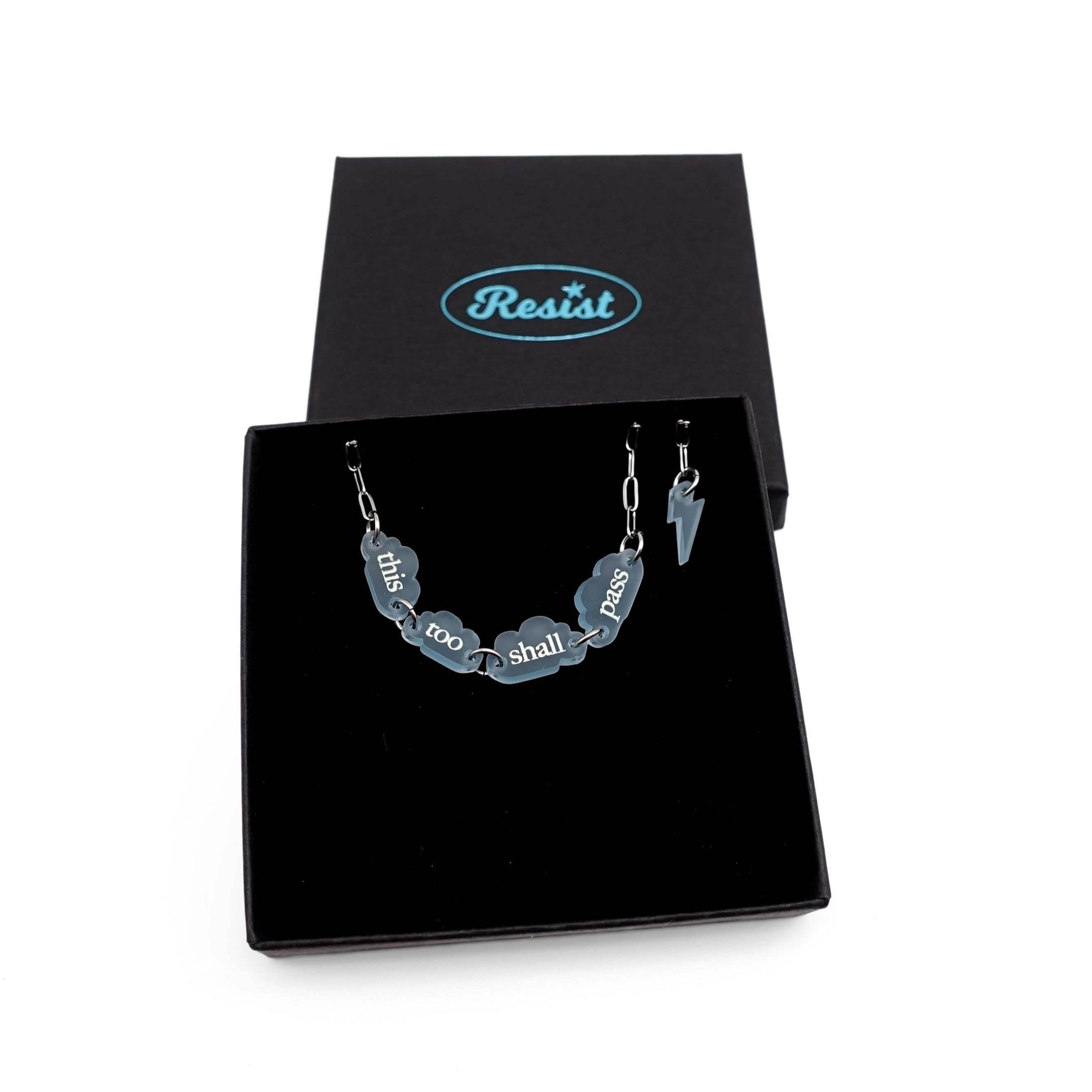 This too shall pass bracelet with words etched in glow-in-the-dark pigment on small clouds on a stainless steel chain, show in a Wear and Resist gift box. 