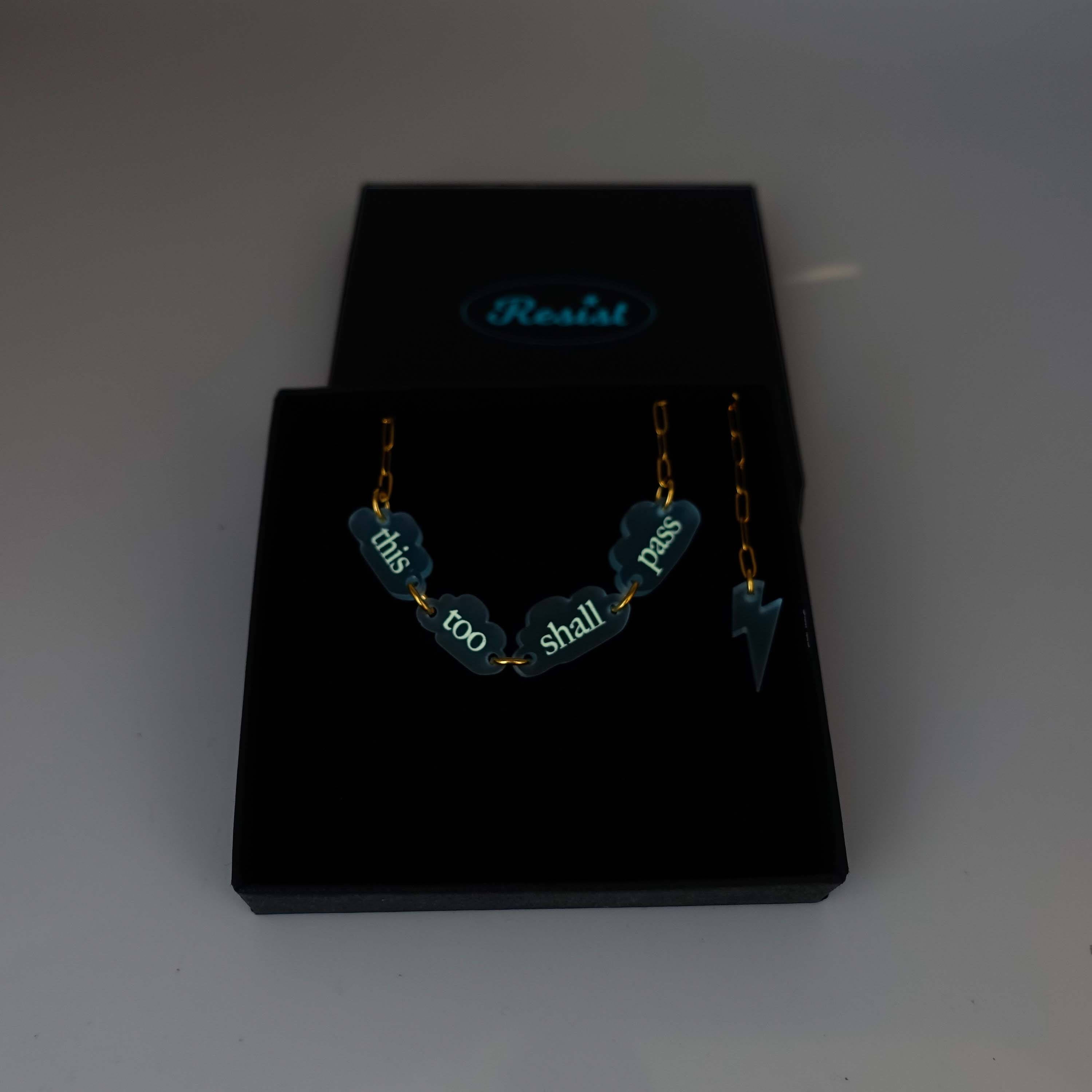 This Too Shall Pass floating clouds necklace shown on an 18K gold ion-plated stainless steel chain shown glowing in the dark in a Wear and Resist gift box.