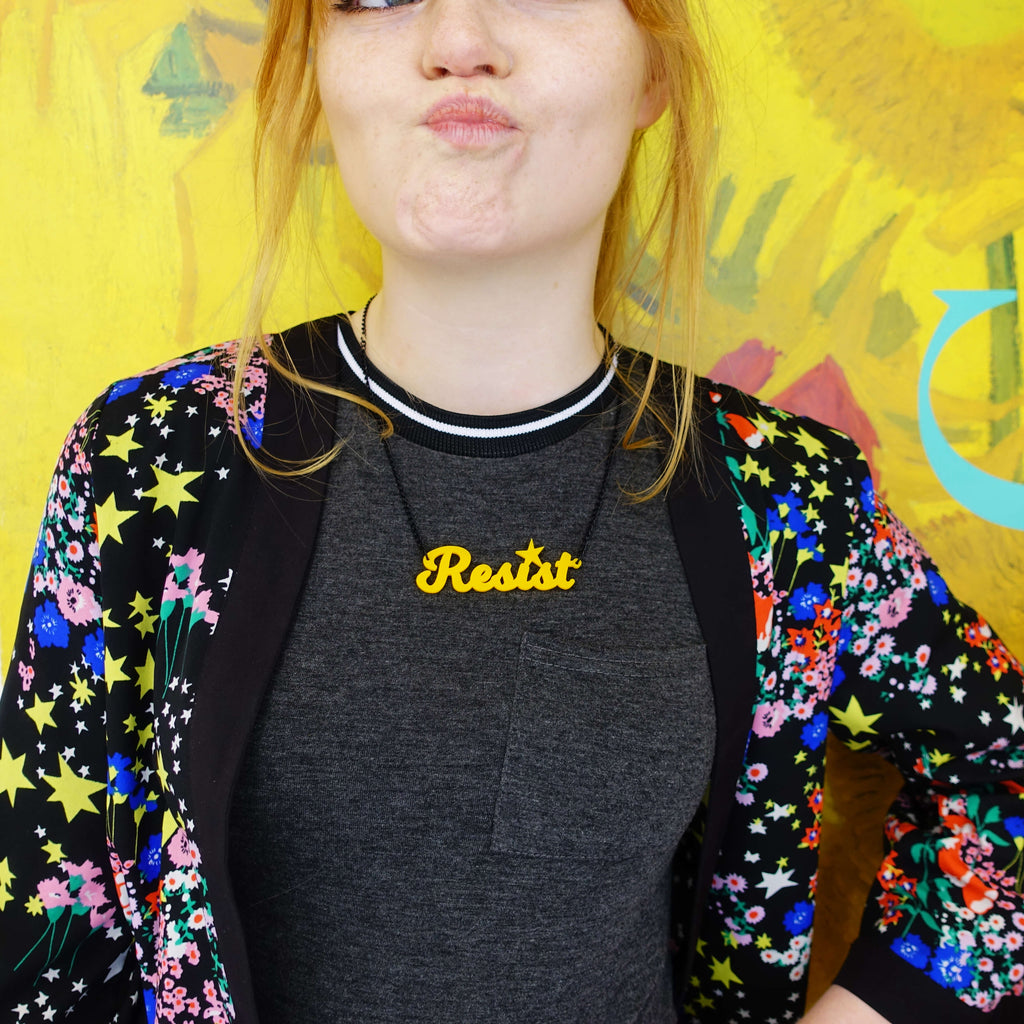 Model wears a sunflower yellow Resist necklace in front of a poster of Van Gogh's sunflowers. Join the Resisterhood! Wear and Resist was started in early 2017 by Sarah Day  to channel her rage into a creative pursuit and raise money for women's charities