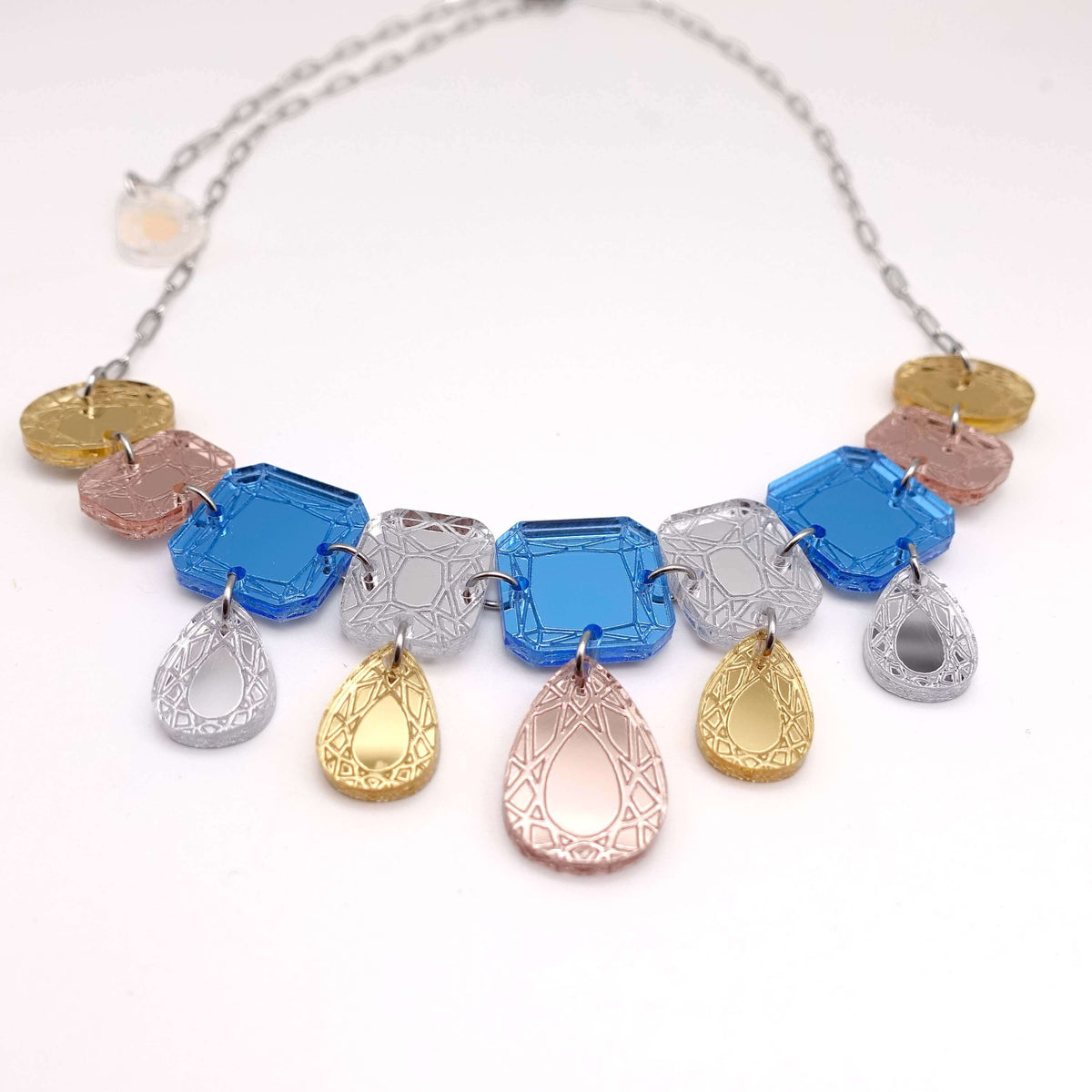 An acrylic necklace made with etched 'jewels' in pale blues, golds and silver, shown on a white surface. 