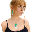 Eliza wears a green Rude Bird pendant necklace that says 'F*ck this' with matching green earrings. 