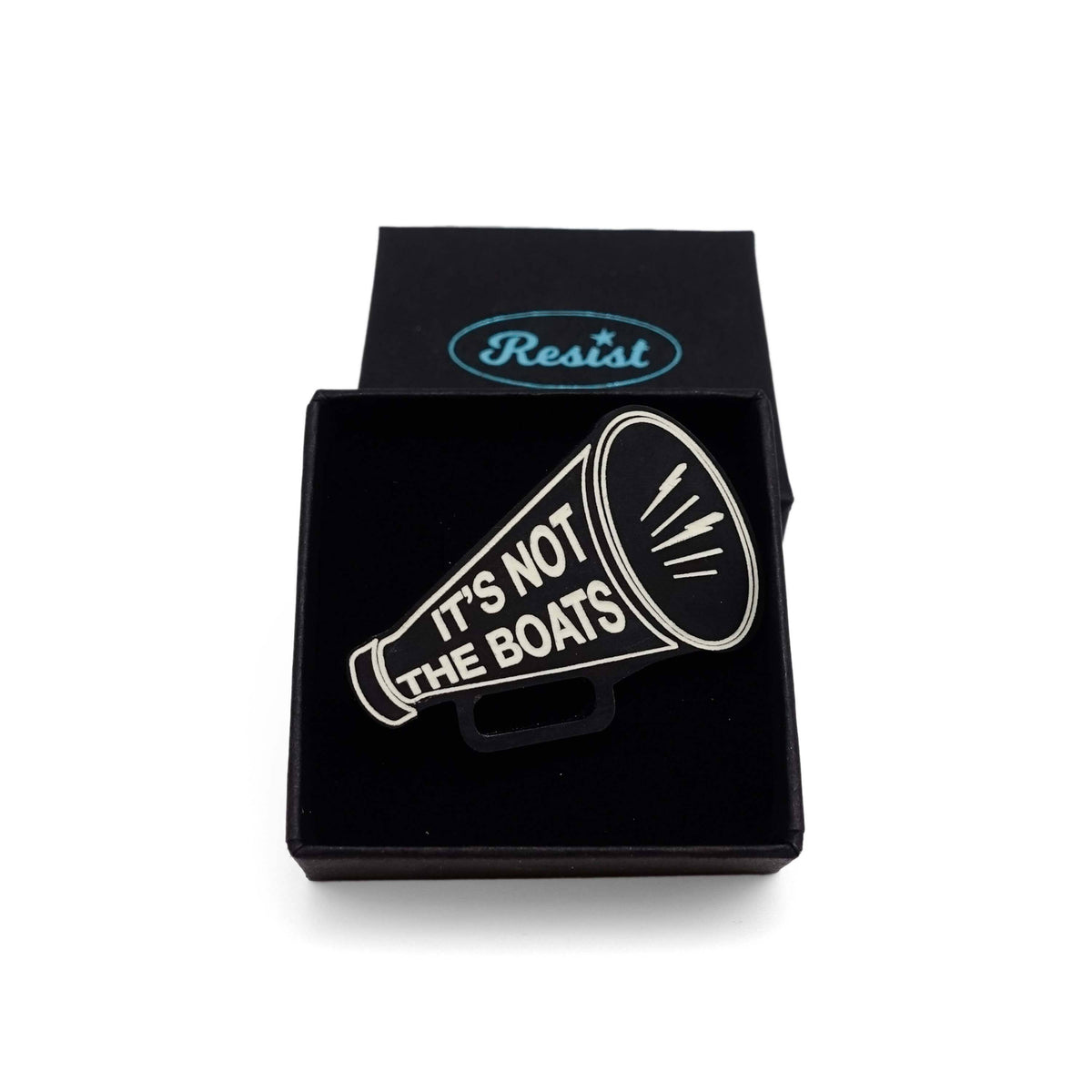 A matte black acrylic brooch in the shape of a megaphone etched in glow-in-the-dark pigment with the words 'It's not the boats', shown in a small Wear & Resist gift box.