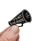 A matte black acrylic brooch in the shape of a megaphone etched in glow-in-the-dark pigment with the words 'It's not the boats', shown held up in my fingers.