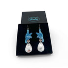 A pair of acrylic earrings with blue birds holding silver 'jewels' that say 'P*ss Off'. Shown in a Wear & Resist gift box. 