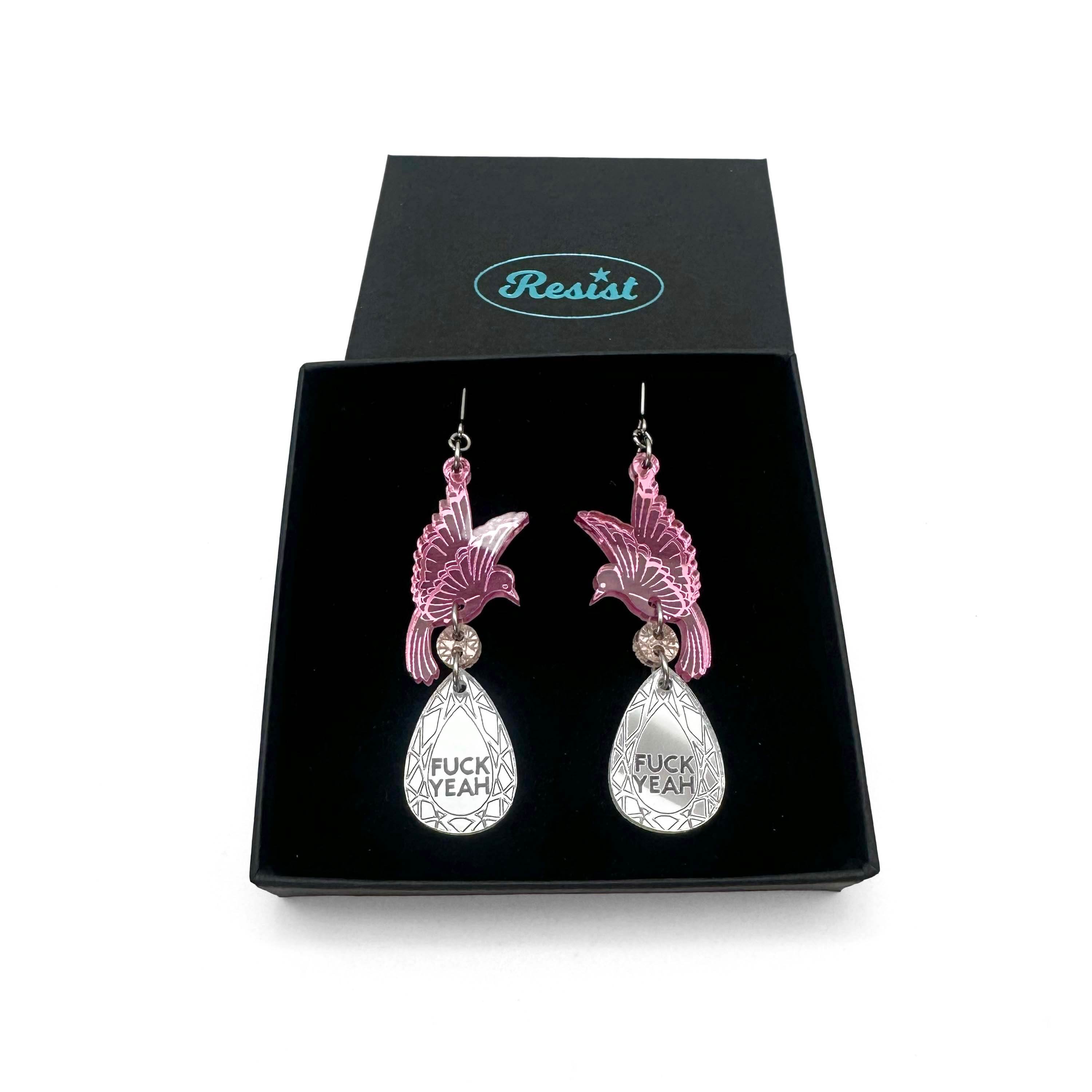 A pair of acrylic earrings with pink birds holding silver 'jewels' that say 'F*ck Yeah'. Shown in a Wear & Resist gift box. 