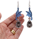 A pair of acrylic earrings with blue birds holding silver 'jewels' that say 'P*ss Off'. Photographed with my hand behind for scale. 