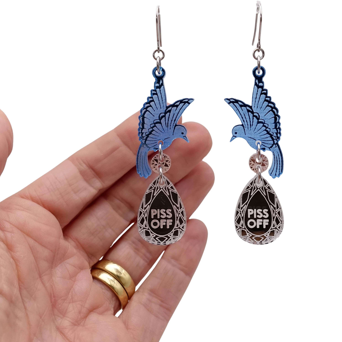 A pair of acrylic earrings with blue birds holding silver 'jewels' that say 'P*ss Off'. Photographed with my hand behind for scale. 
