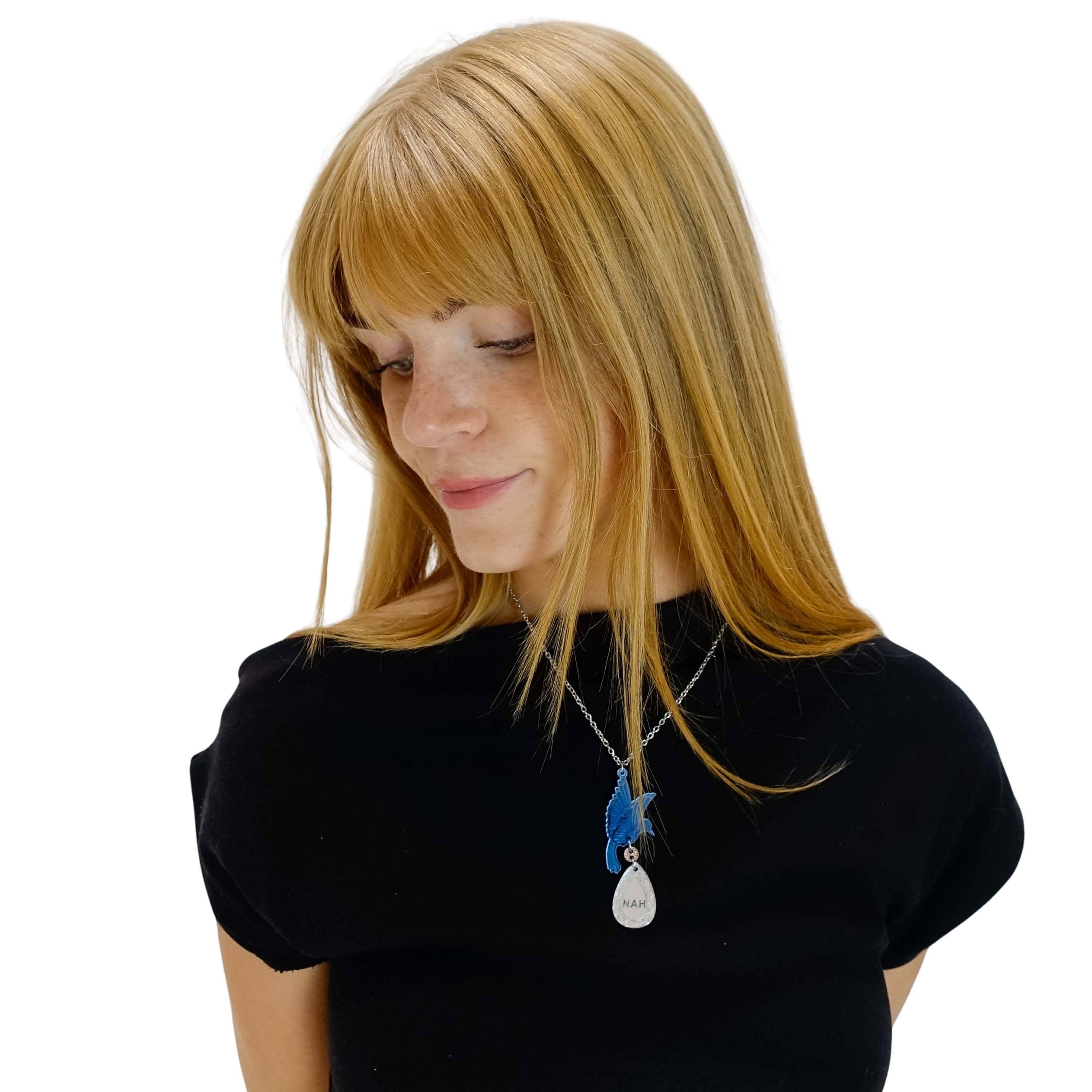 Eliza wears a Rude Bird acrylic pendant necklace with a blue bird holding a silver 'jewel' that says 'Nah'.  
