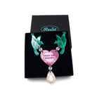 A mirror acrylic necklace with two green birds holding a pink heart that says 'Fuck the Fascists' with a rose gold 'jewel' hanging from the centre, shown in a Wear and Resist gift box. 