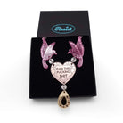 A mirror acrylic necklace with two pink birds holding a rose gold heart that says 'Fuck this fucking shit' with a gold 'jewel' hanging from the centre. Shown in a Wear and Resist gift box. 