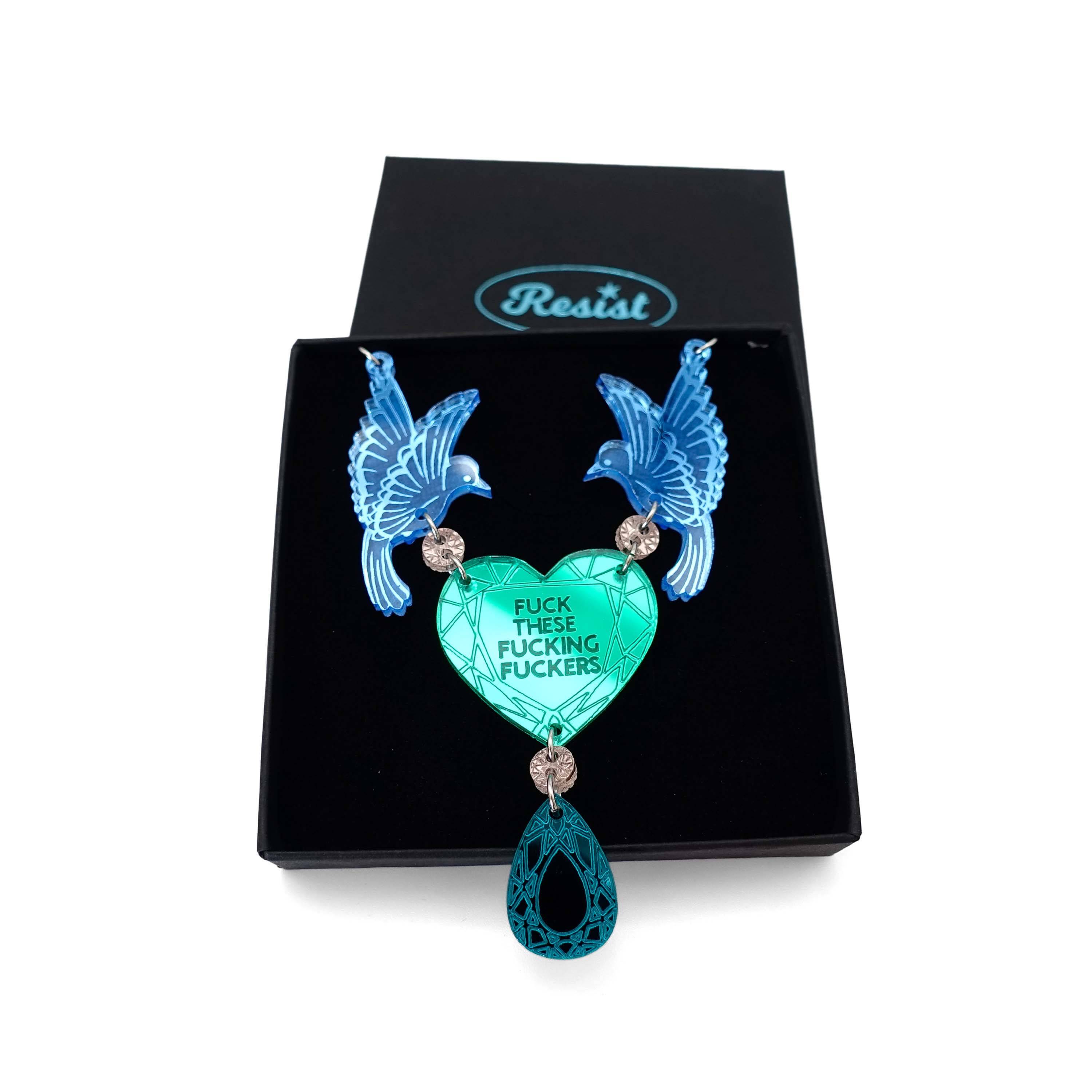 A mirror acrylic necklace with two blue birds holding a green heart that says 'Fuck these fucking fuckers' with a teal gold 'jewel' hanging from the centre. Shown in a Wear and Resist gift box. 