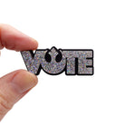 A silver glitter VOTE brooch with Rebel Alliance symbol from Star Wars, shown held up for scale. 