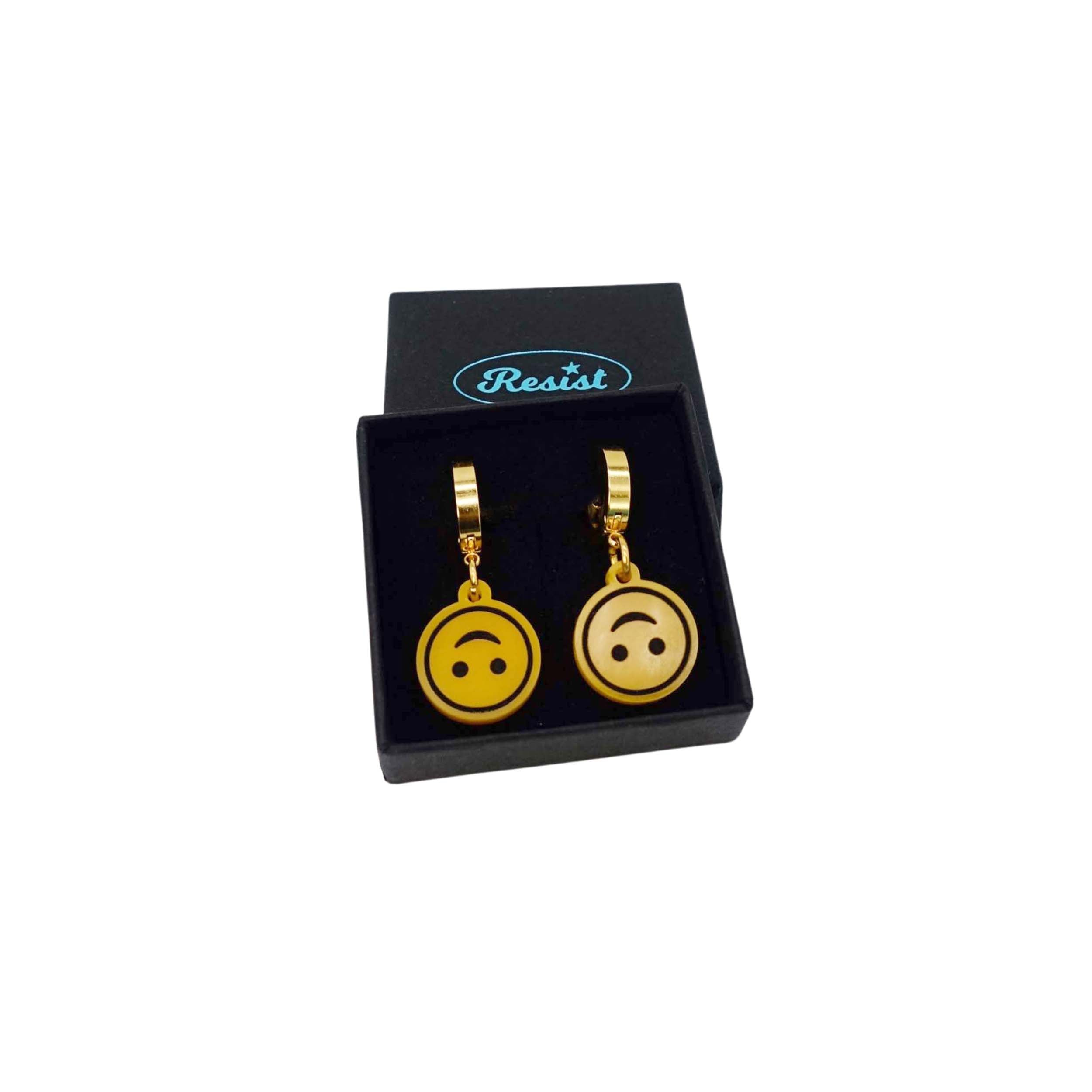 Upside-down smiley face emoji earrings on gold huggie hoops shown in a Wear and Resist gift box. 
