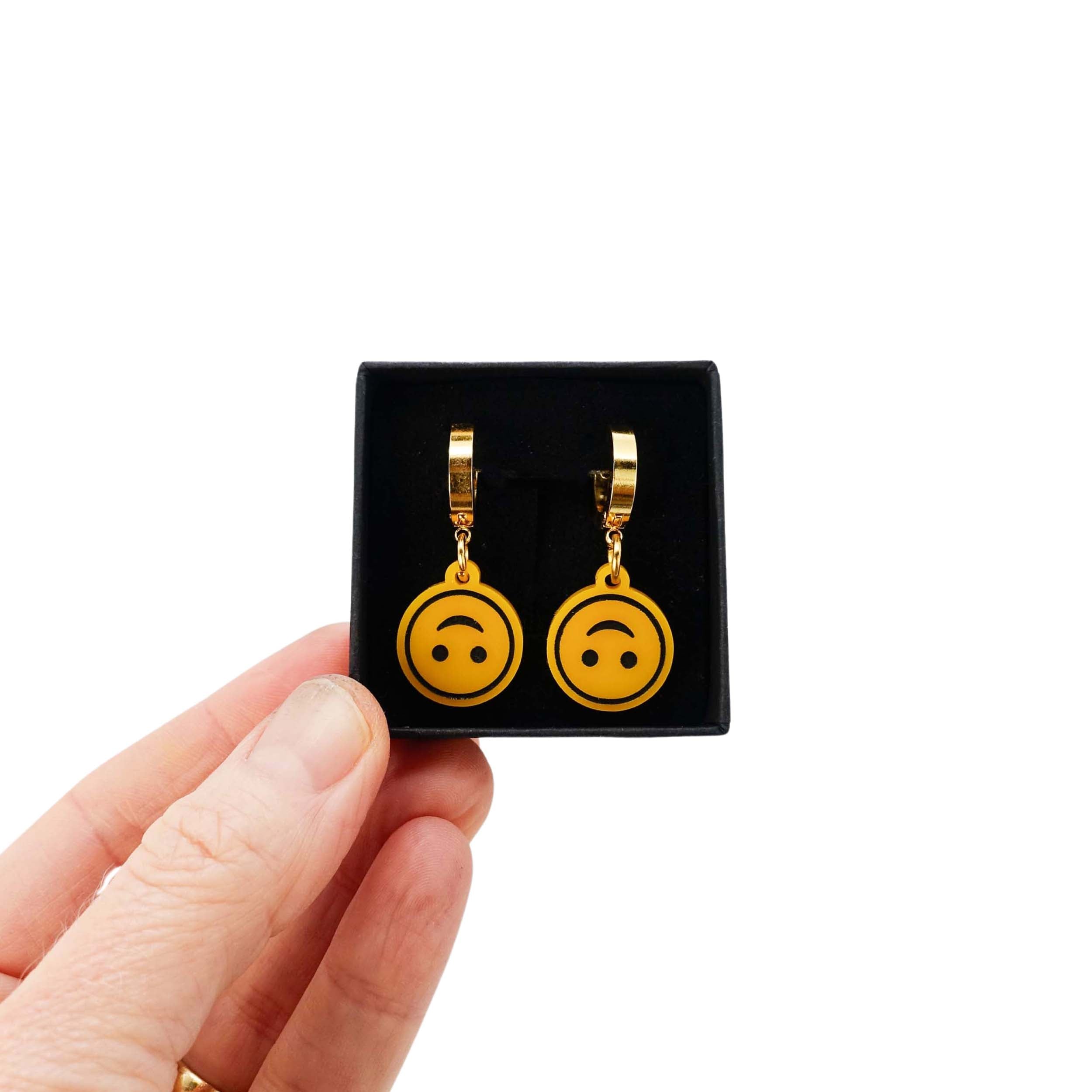 Upside-down smiley face earrings on gold  huggie hoops, shown held up in a Wear and Resist gift box.
