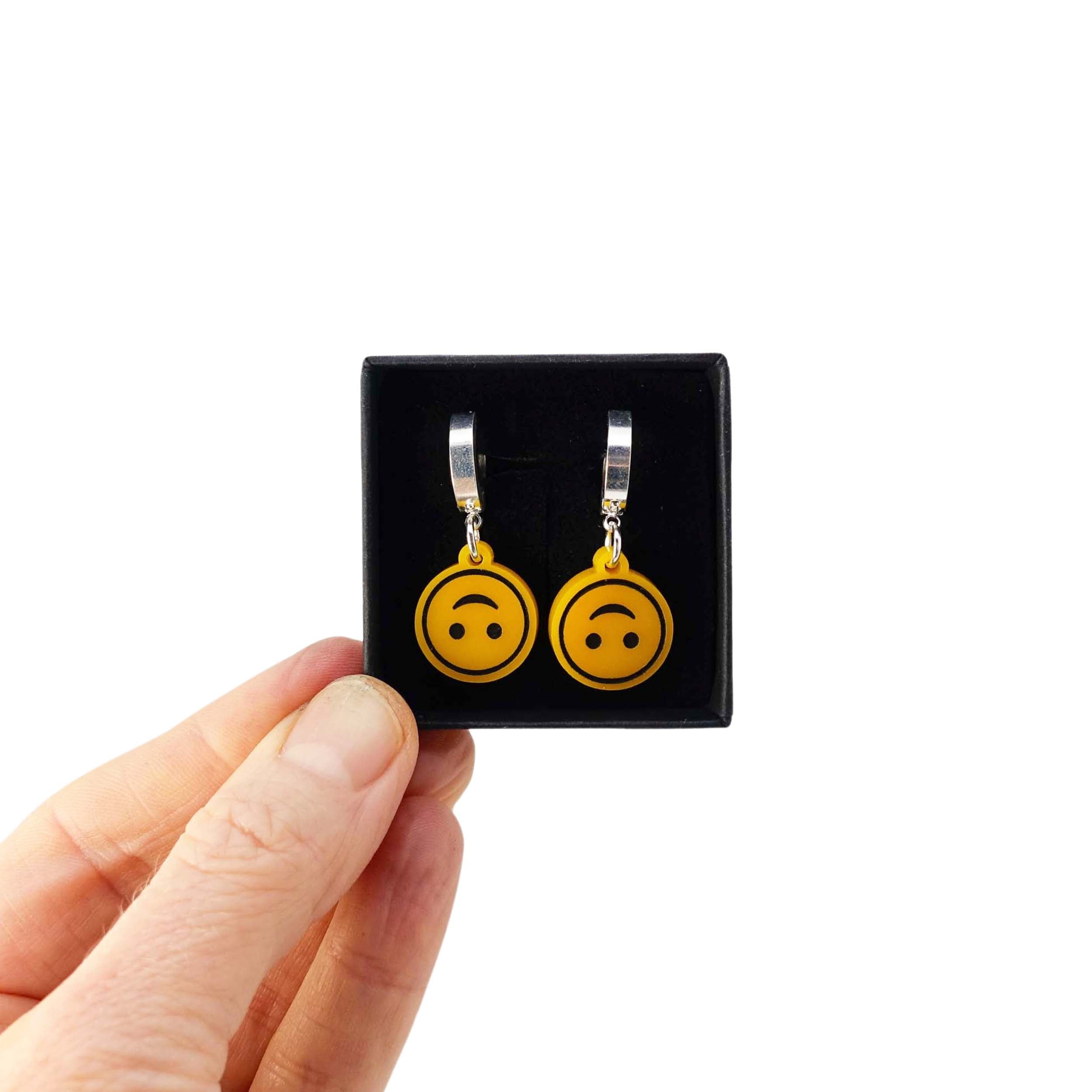 Upside-down smiley face earrings on silver huggie hoops, shown held up in a Wear and Resist gift box.