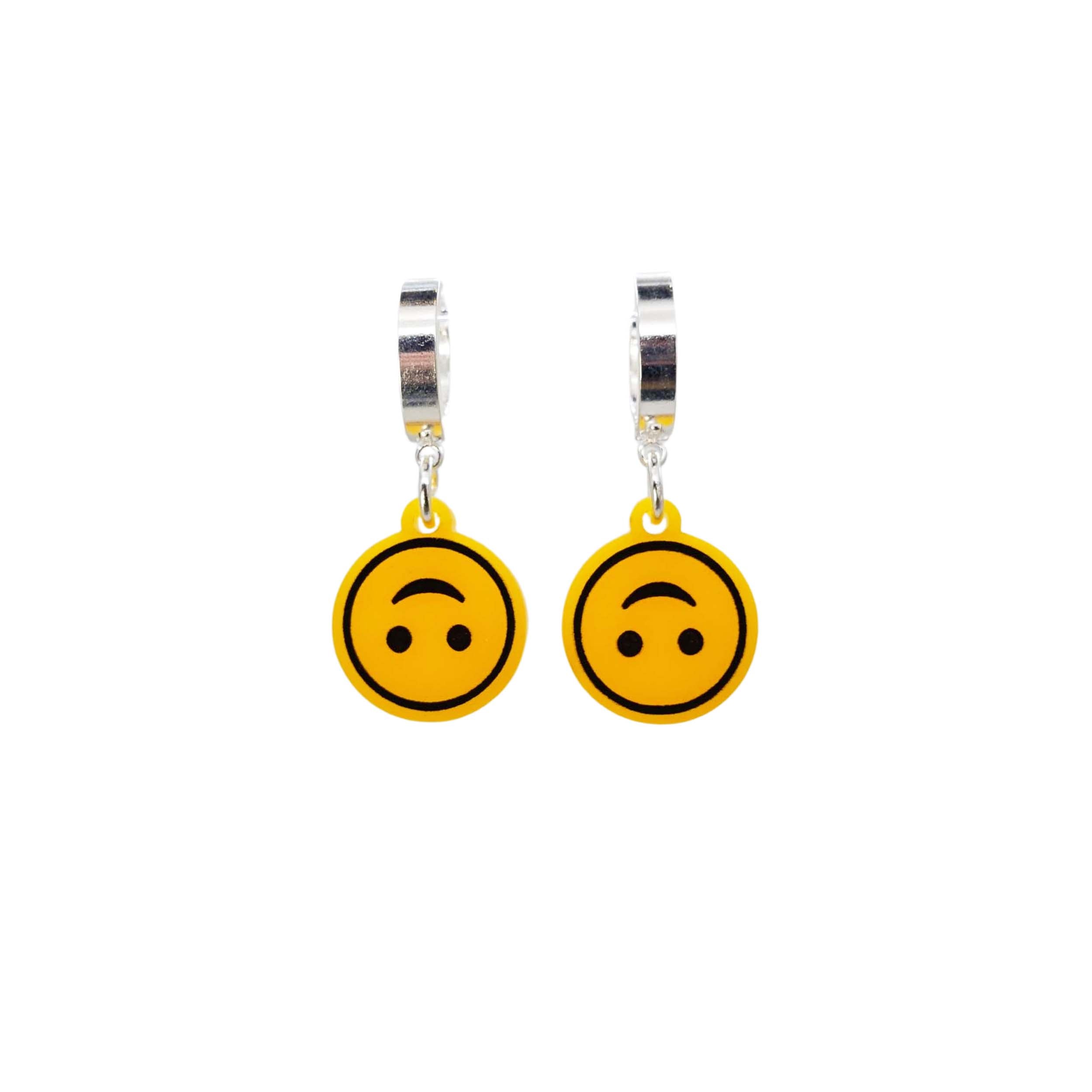 Upside-down smiley face emoji earrings on silver huggie hoops, shown hanging against a white background. 