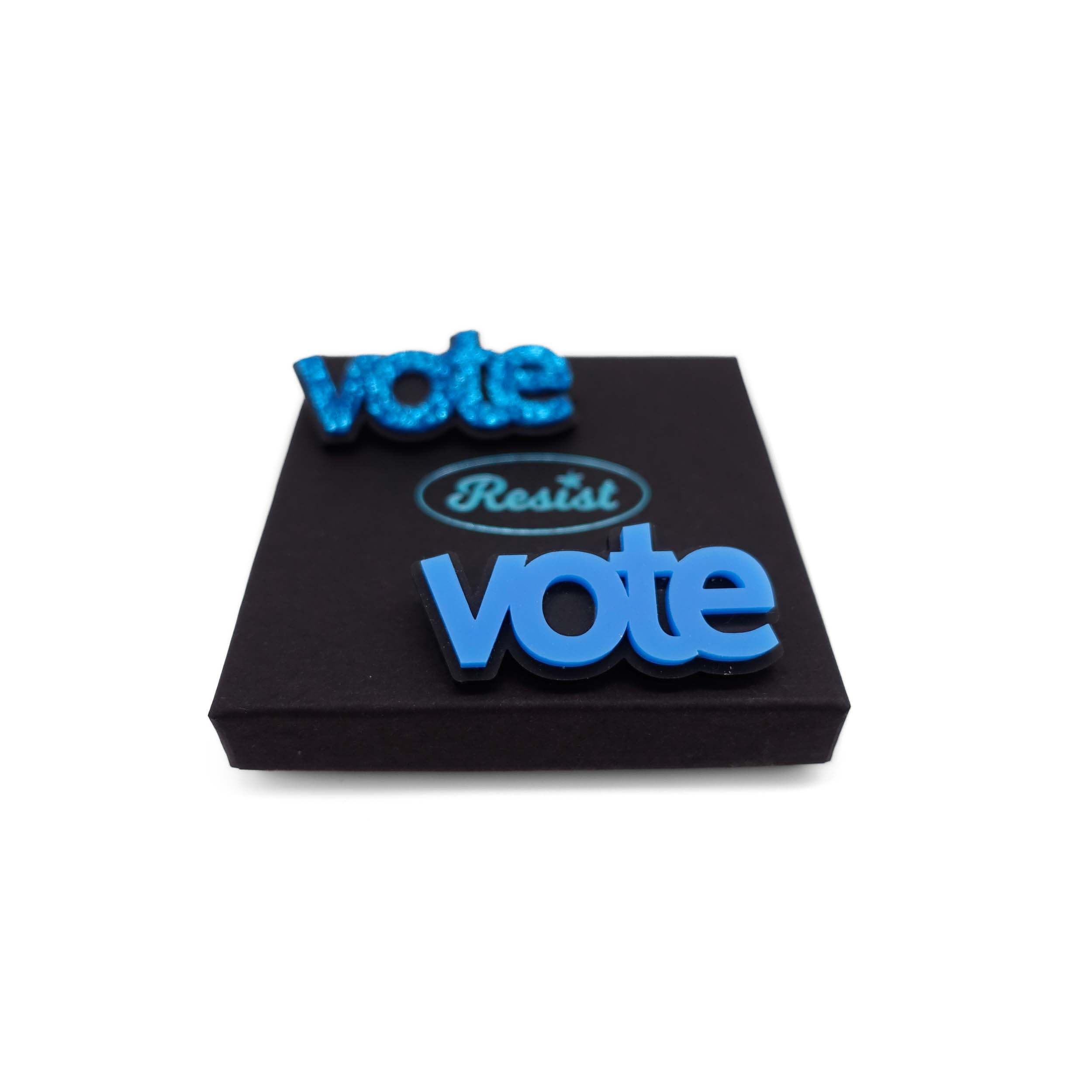 Two Vote brooches in blue glitter and bright matte blue shown on a Wear and Resist gift box. Get any two items in the Vote Blue Collection for £36 with code VOTEBLUE.