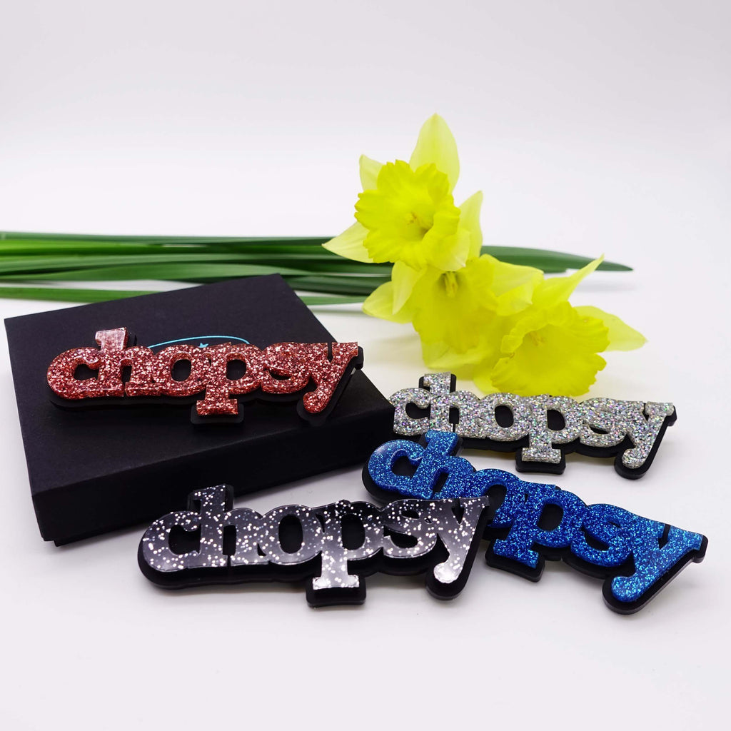 All four glitter colours of Chopsy brooches shown with a Wear and Resist gift box and daffodils. £2 goes to Welsh Women's Aid. Fel yr arfer, mae £2 o bob mwclus Chopsy yn mynd at @welshwomensaid.