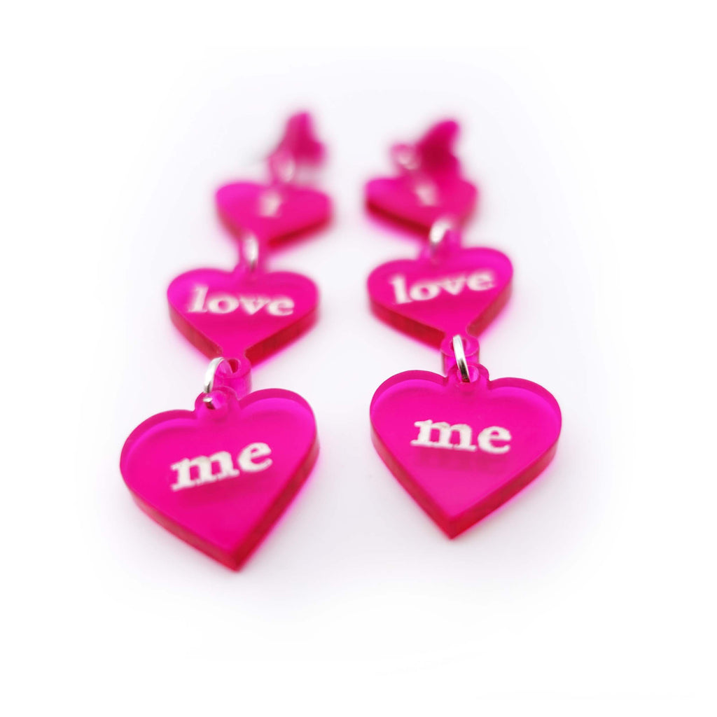 I love me Valentine's earrings designed by Sarah Day for Wear and Resist, because self-love is the best love. Three clear acrylic fuschia hearts are engraved with the words. 