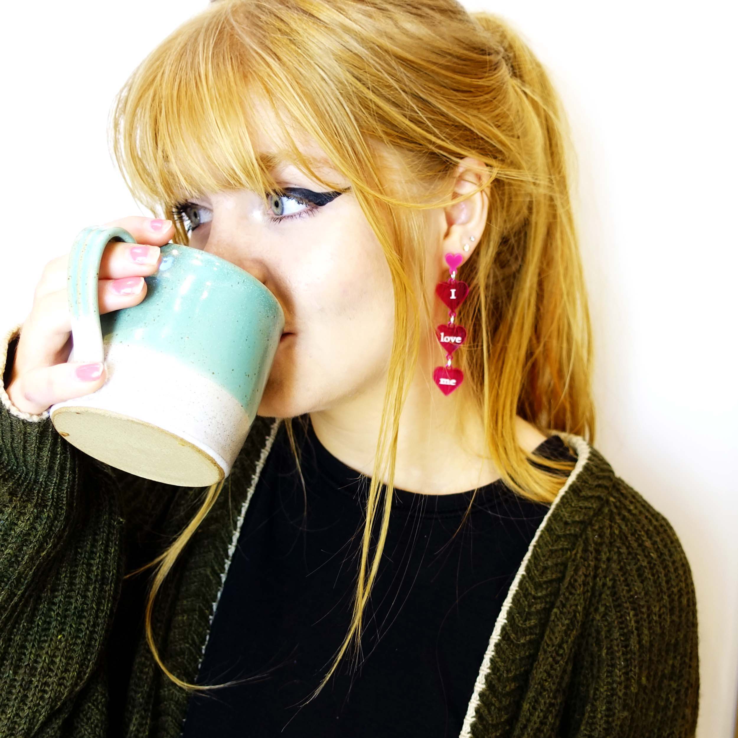 Eliza drinks a cup of tea wearing I love me earrings by Wear and Resist. 