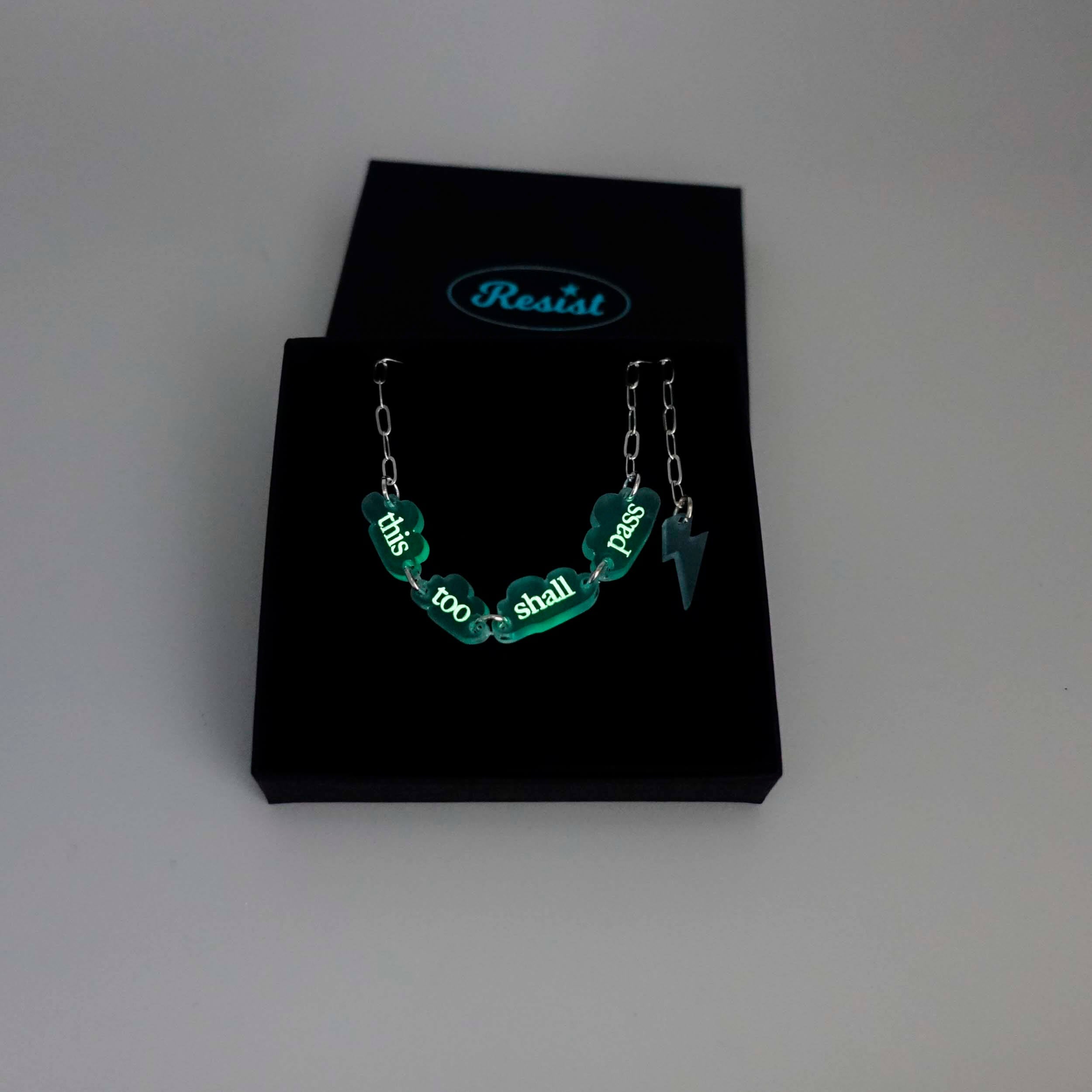 This too shall pass floating clouds necklace on a  silver-plated paper-clip chain with lightning bolt charm. The text is etched with glow-in-the-dark pigment and is shown in half-light glowing in a Wear and Resist gift box. 