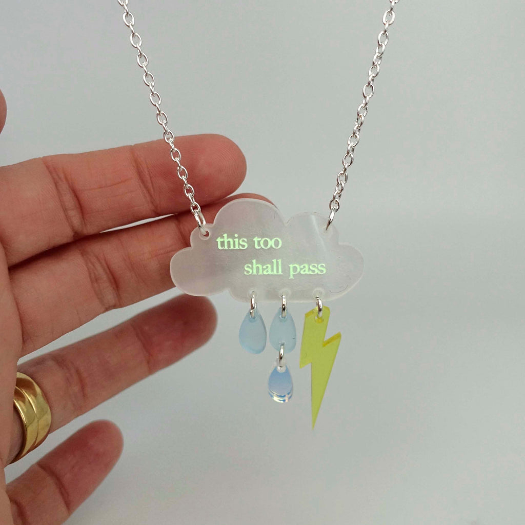 This too shall pass raincloud pendant necklace shown in half-light with the glow in the dark pigment beginning to glow.