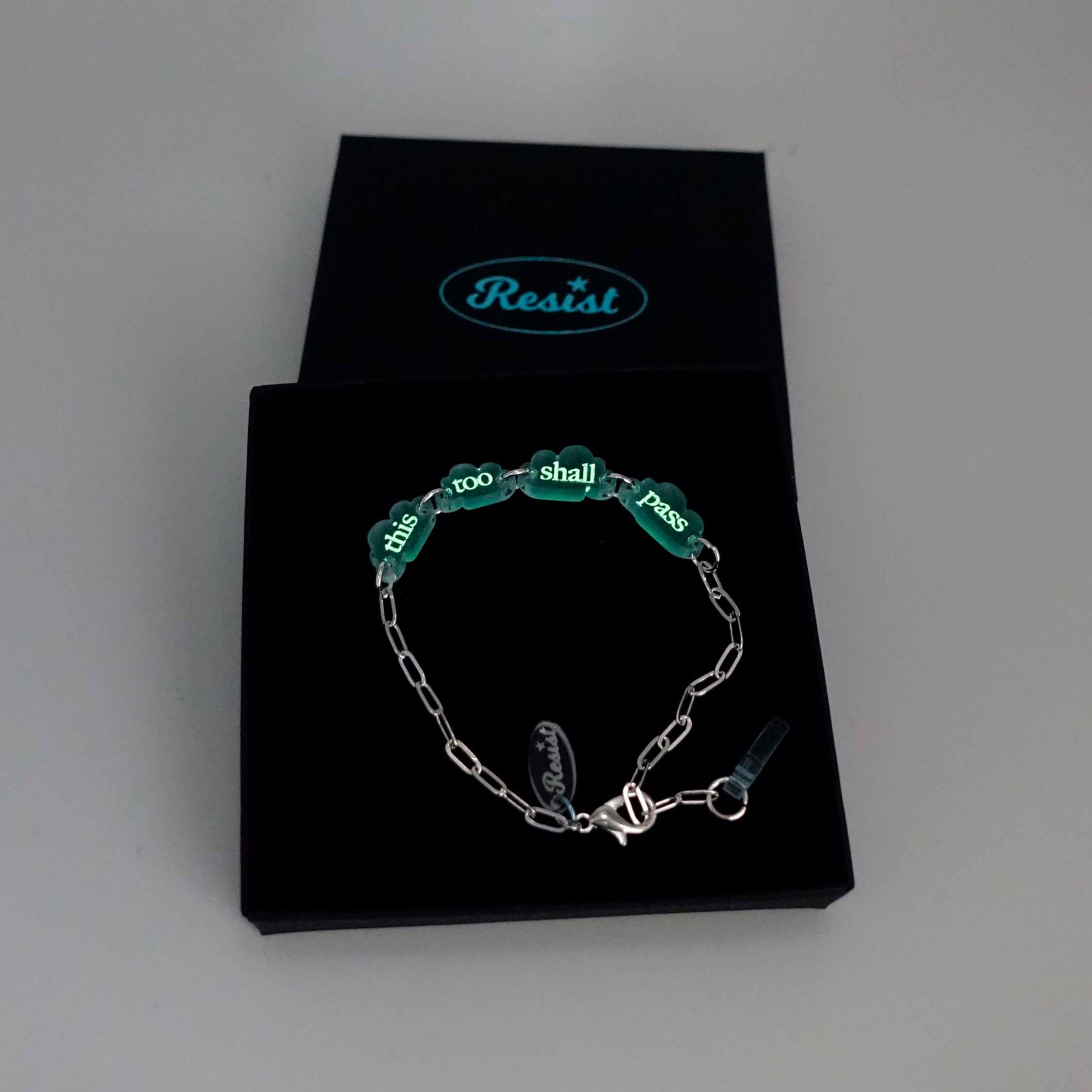 This too shall pass floating clouds silver-plated paperclip bracelet, etched with glow-in-the-dark pigment shown in a Wear and Resist gift box. 