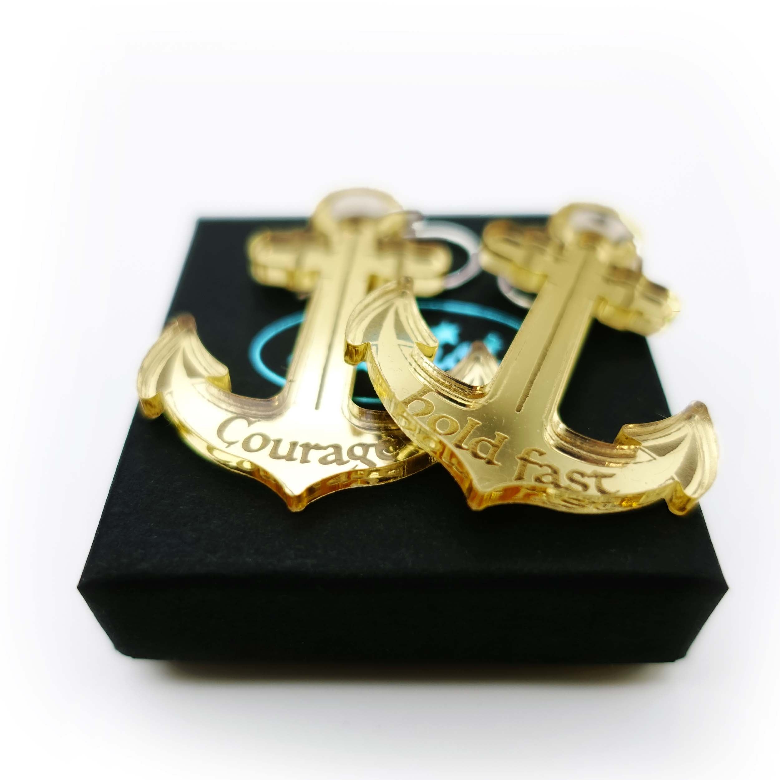 Pair of mirror gold anchor earrings etched with the words courage and hold fast on a Wear and Resist gift box in aid of the RNLI and Women for Refugee Women. 